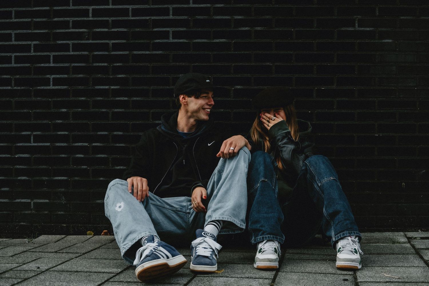 Happy young couple sitting together against a dark brick wall, laughing and smiling outdoors.