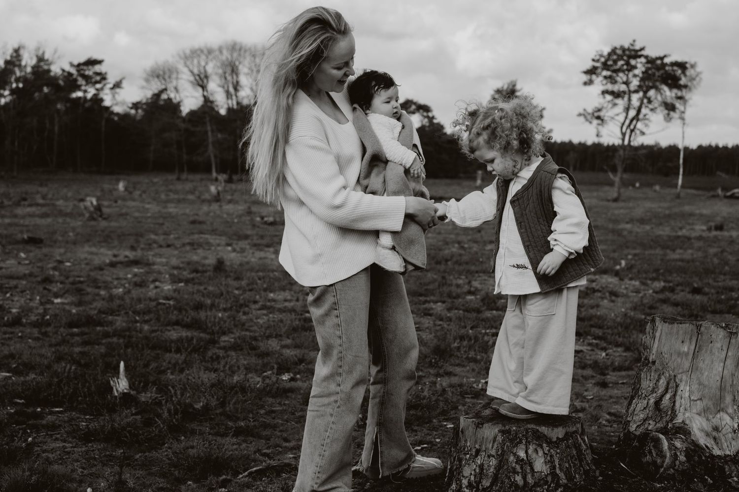 A blonde mother tends to her two young children outdoors in a woodland field.