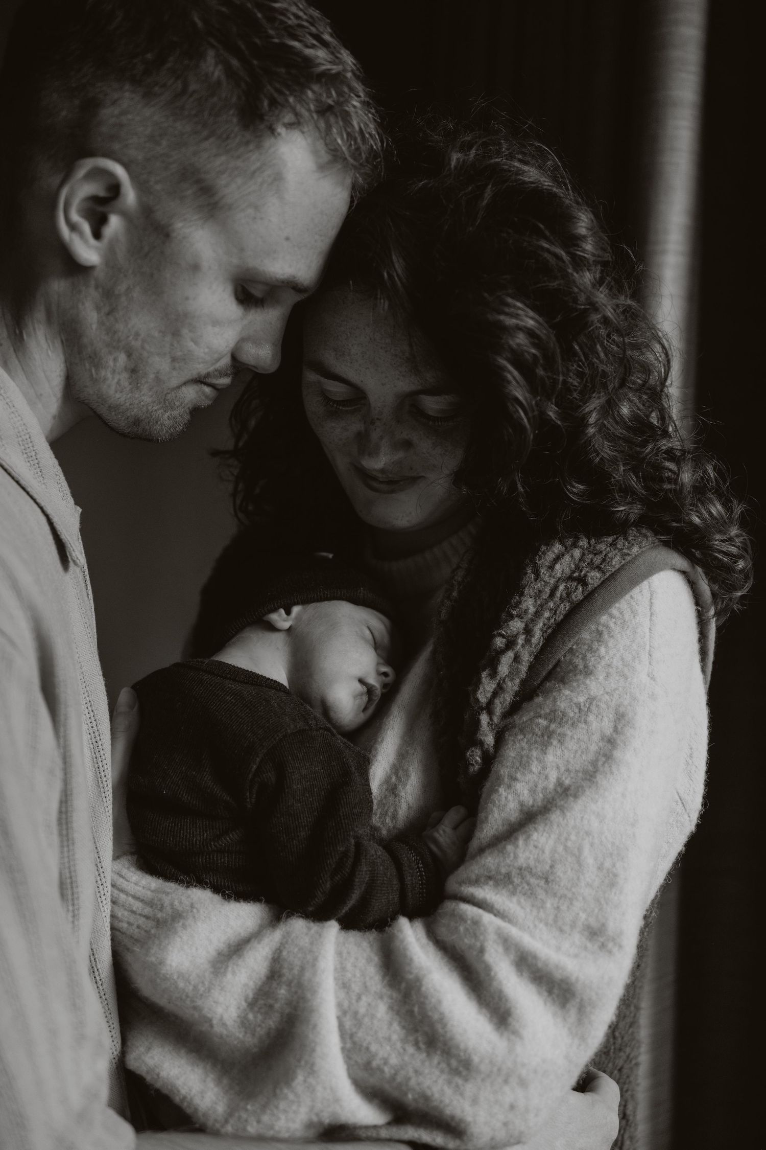 Black and white portrait of new parents tenderly cradling their sleeping newborn baby together.