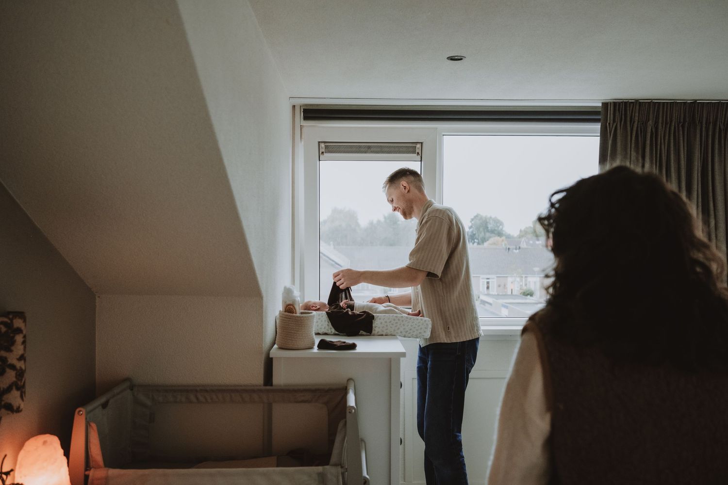 Father prepares baby items at changing table near window while mother watches in nursery room.