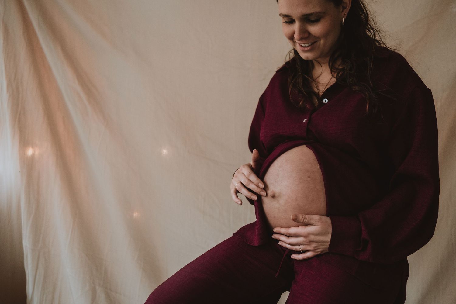 A smiling pregnant woman in burgundy outfit cradles her bare baby bump indoors.