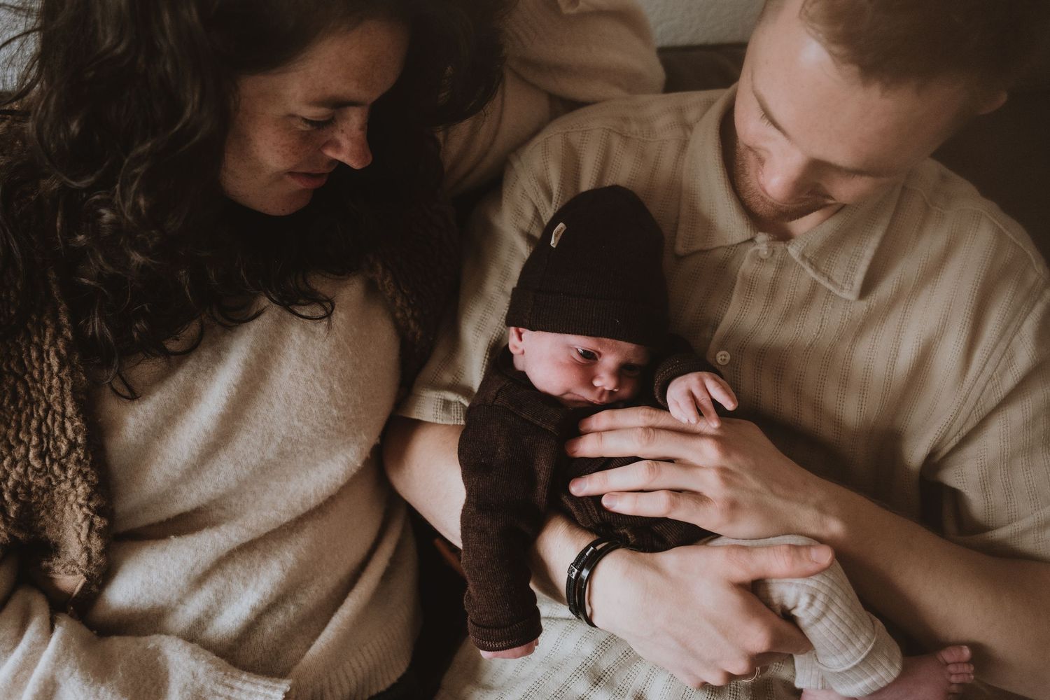 New parents tenderly gaze at their sleeping newborn baby dressed in dark outfit and knit hat.