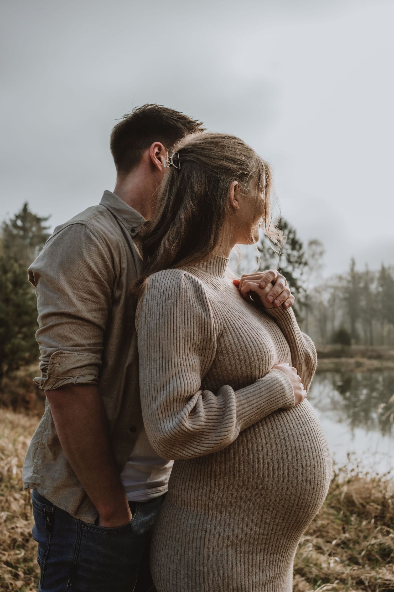 Couple standing close near misty lake, pregnant woman in beige knit dress holding belly, man embracing her.