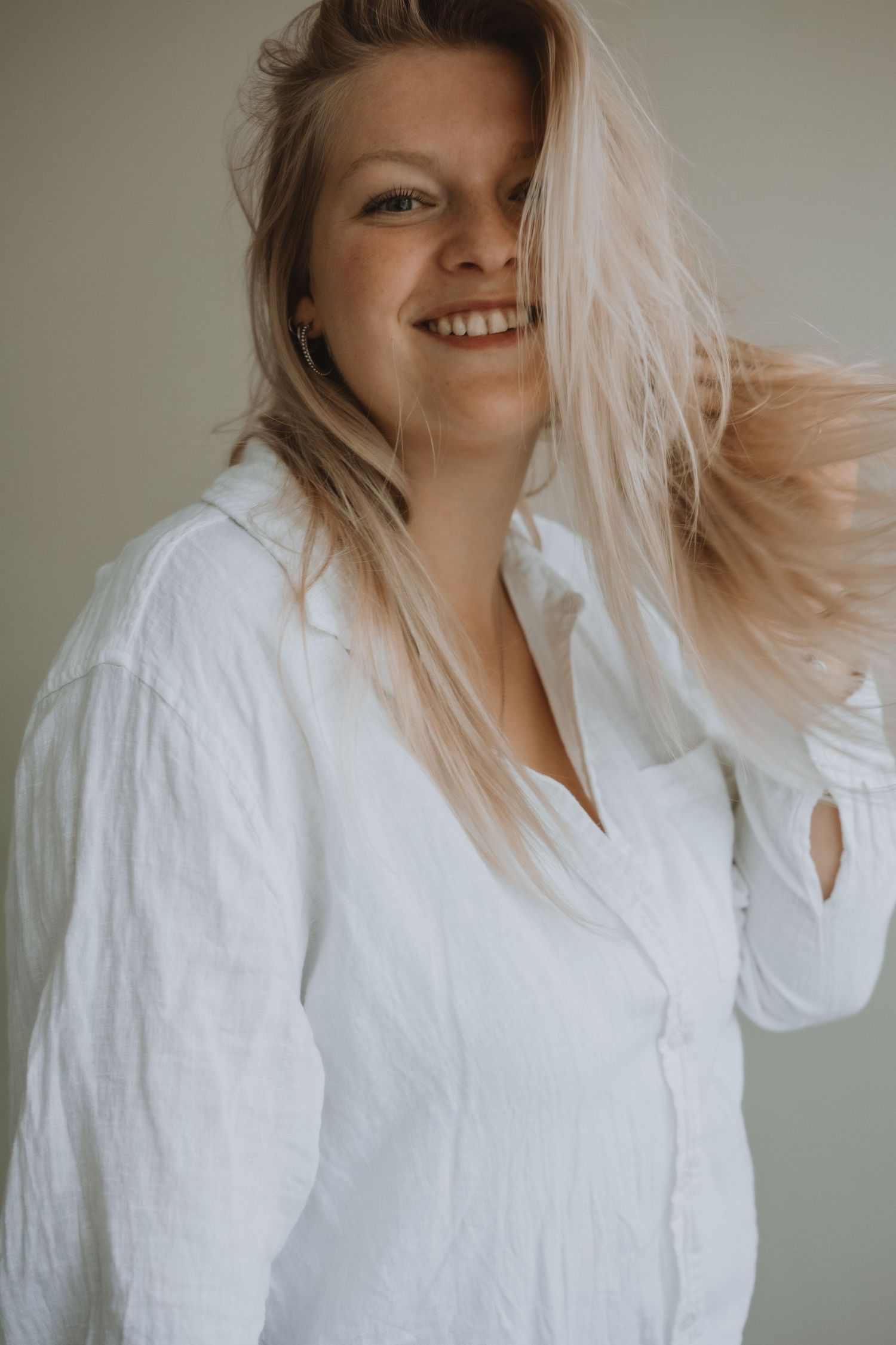 Joyful blonde woman in white linen shirt tosses hair while smiling brightly.