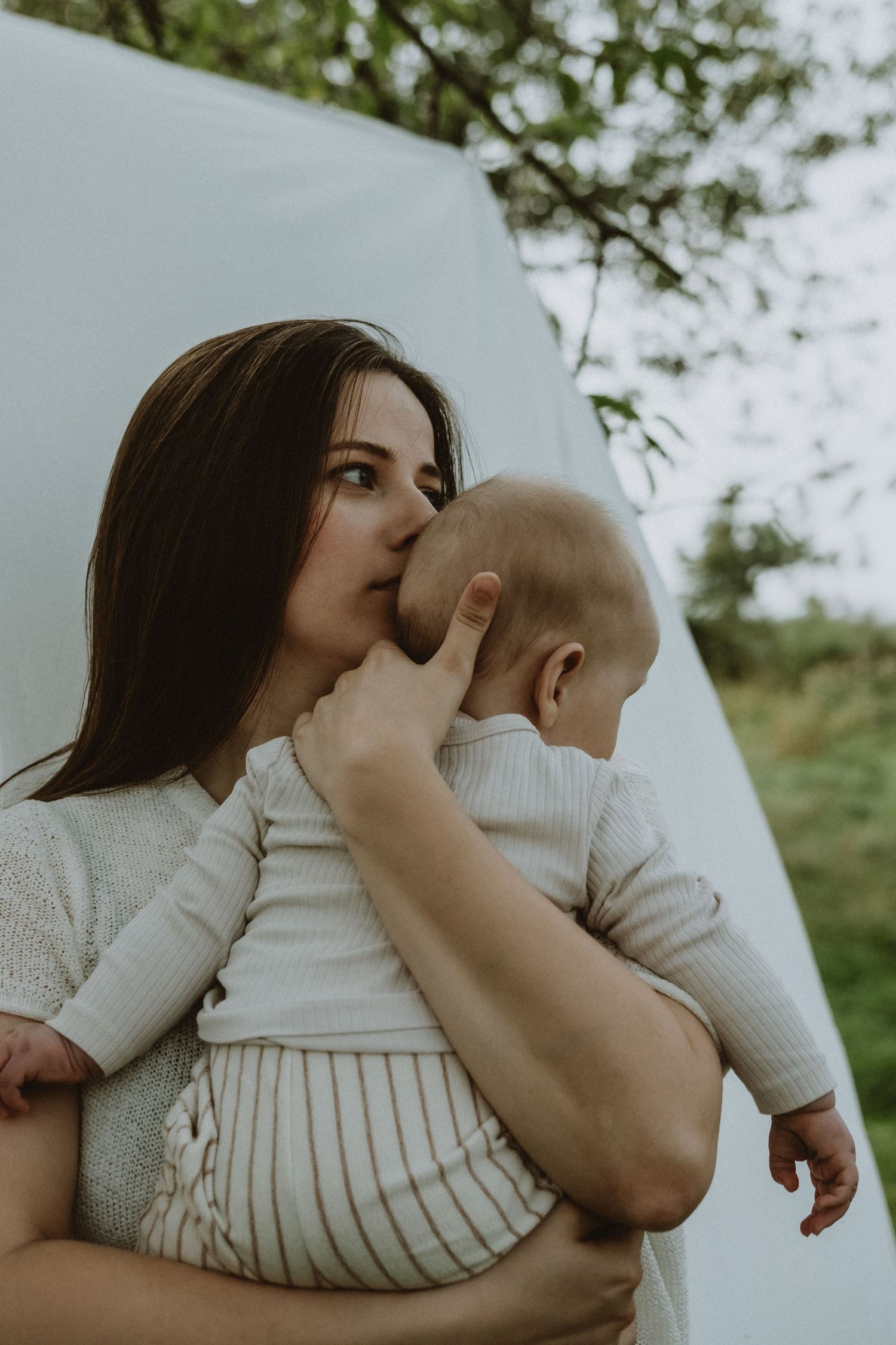 Mother tenderly kissing baby on head while holding them against white backdrop outdoors.