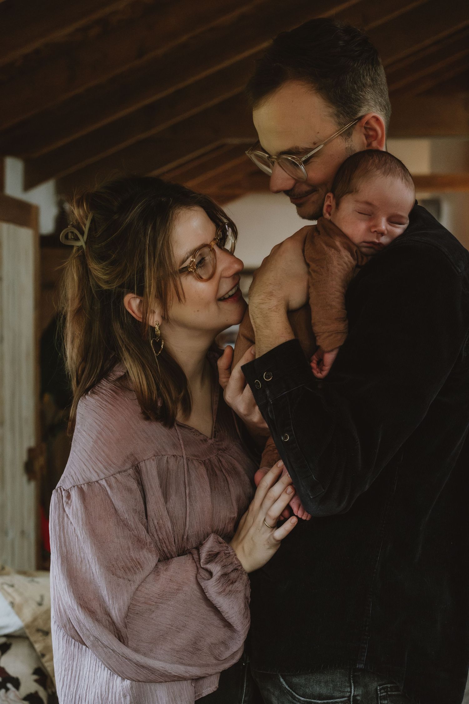 New parents with glasses smiling adoringly at their newborn baby held by father indoors.