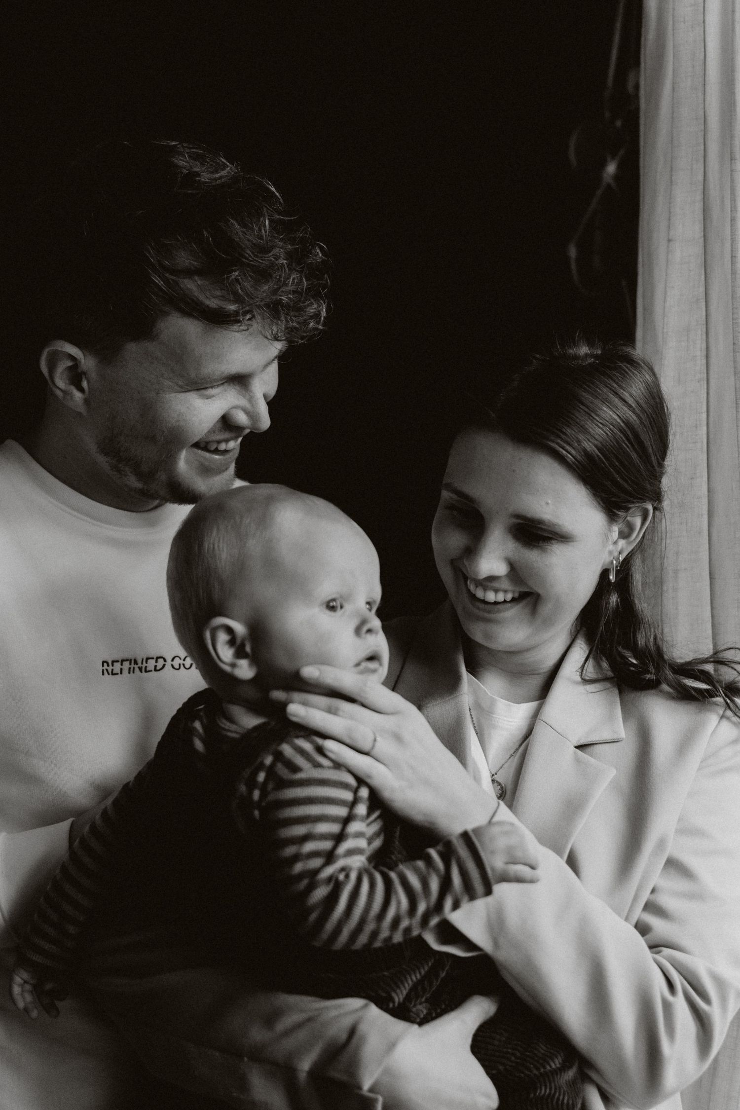 Black and white portrait of smiling parents holding their baby near a window.