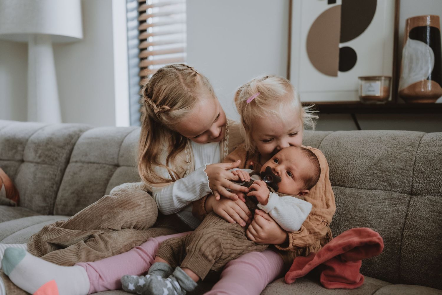 Two young sisters lovingly cuddle and kiss their newborn baby sibling on a gray sofa.