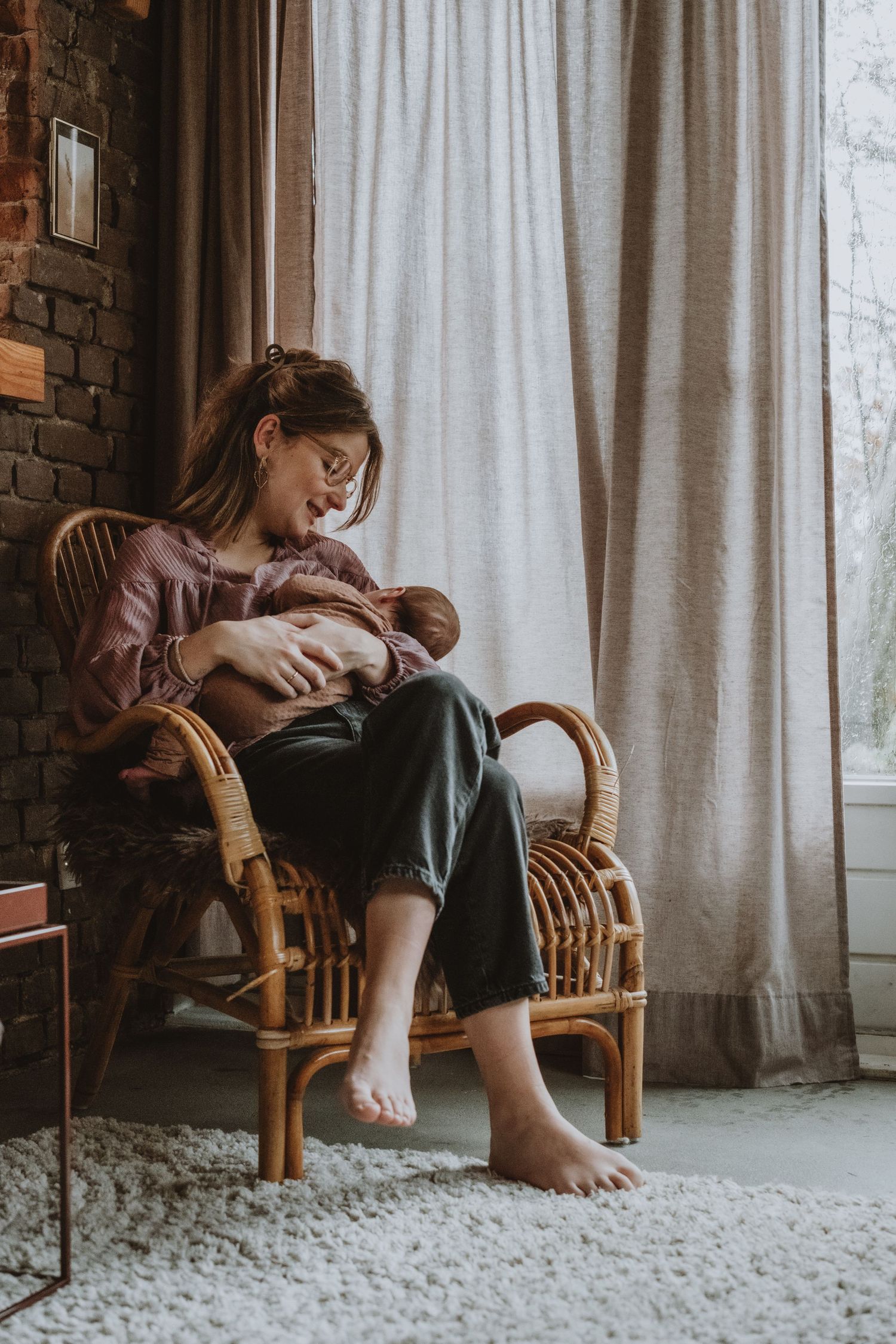 A barefoot mother sits in a rattan chair by a curtained window, lovingly cradling her newborn baby.