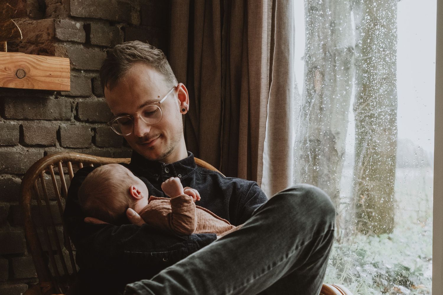 Father with glasses sitting in chair by window tenderly holding sleeping newborn baby.