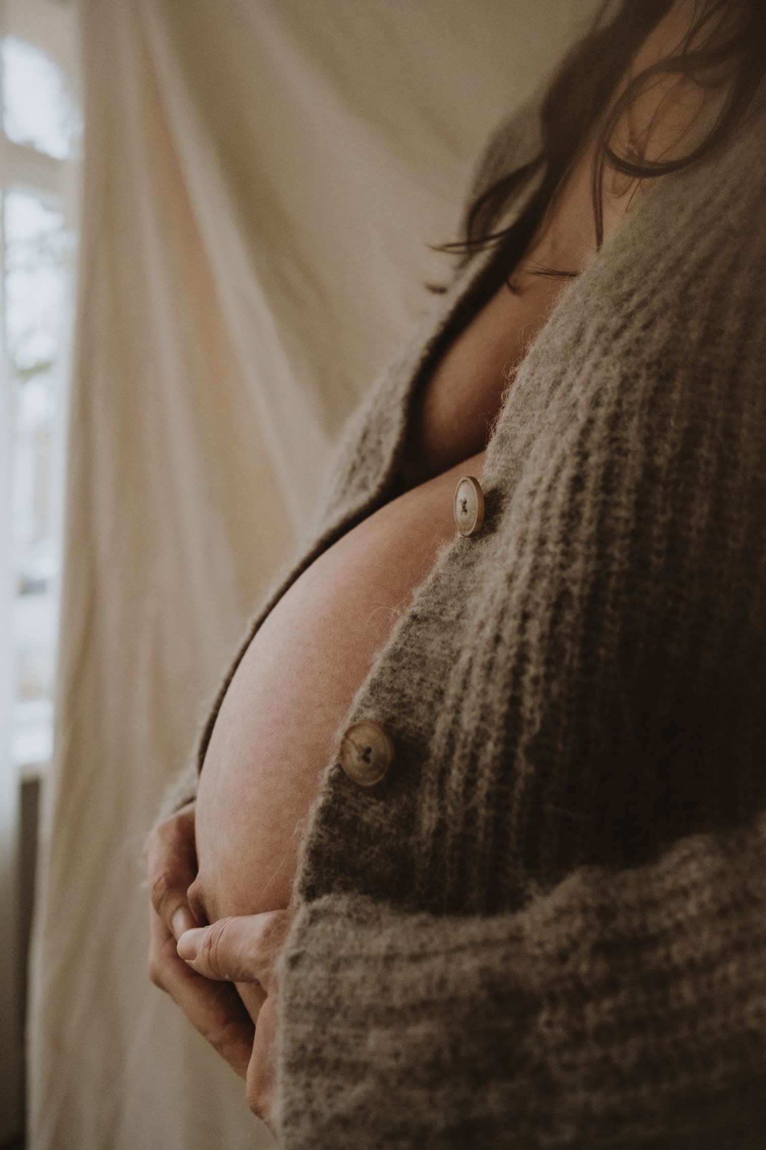Close-up of pregnant woman in gray cardigan gently holding bare baby bump indoors.