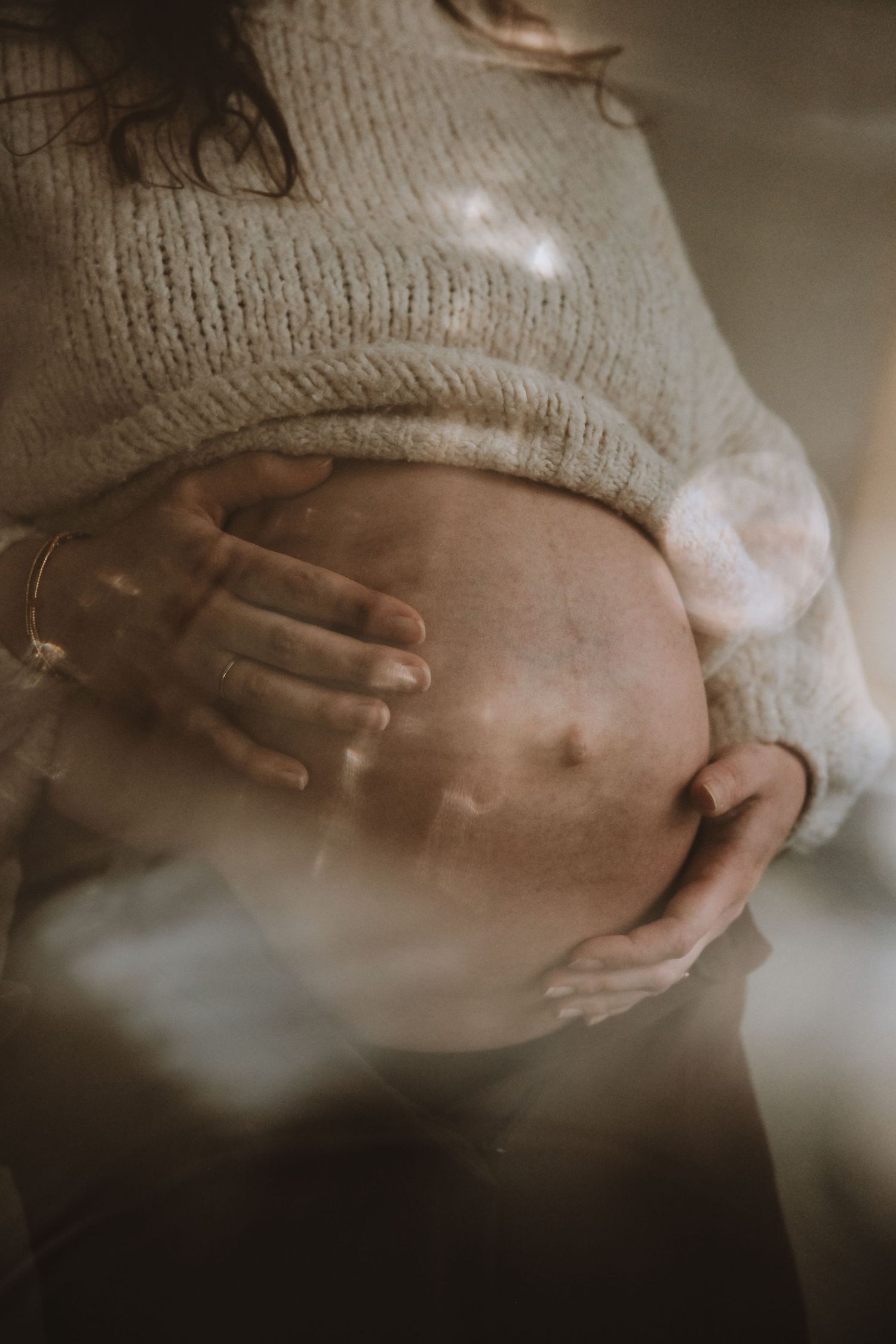 Artistic maternity photo with dreamy light bokeh of hands cradling pregnant belly in knit top.