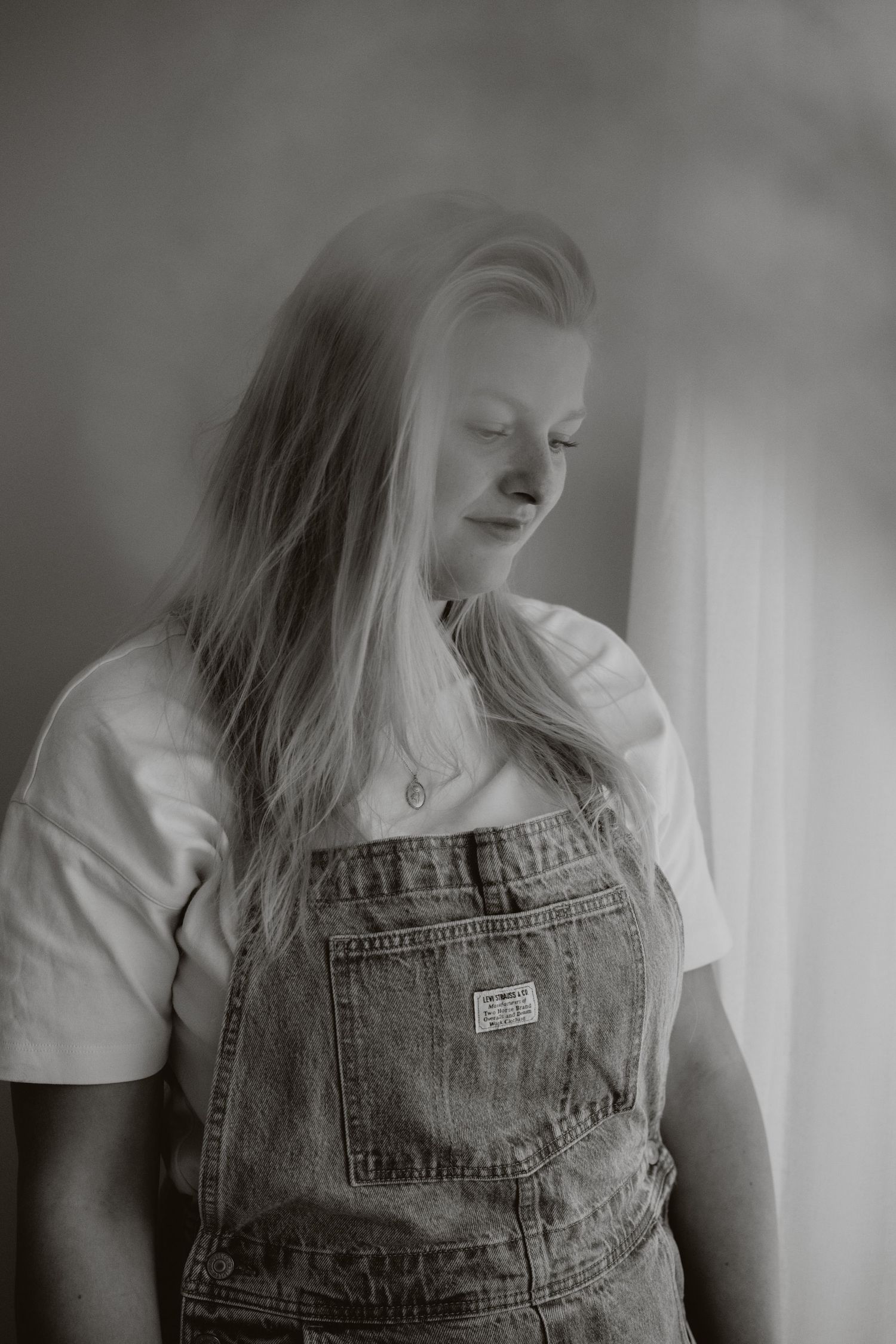 Black and white portrait of young woman in denim overalls standing by a curtained window.
