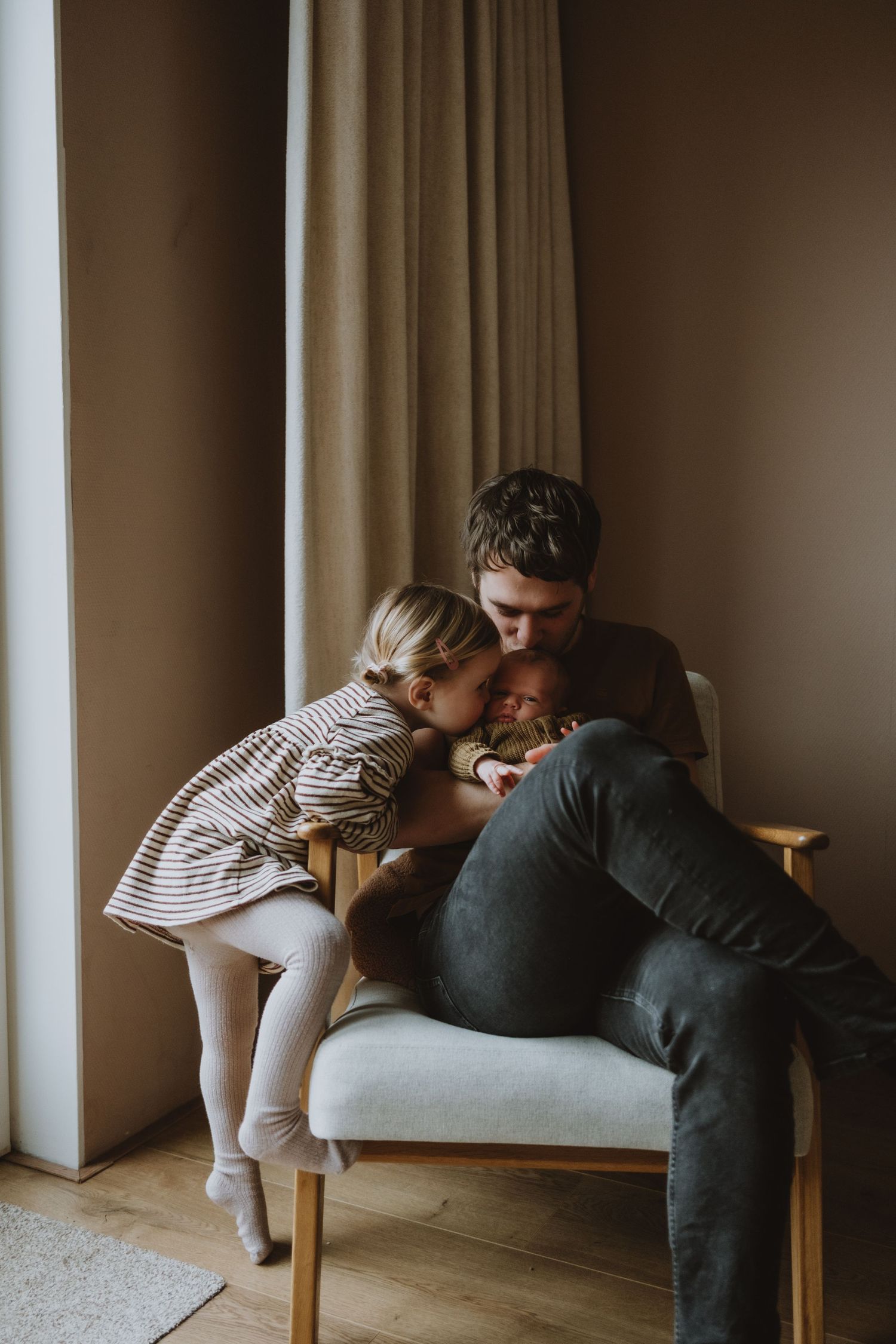 Father holding newborn while child leans in for a kiss in armchair.