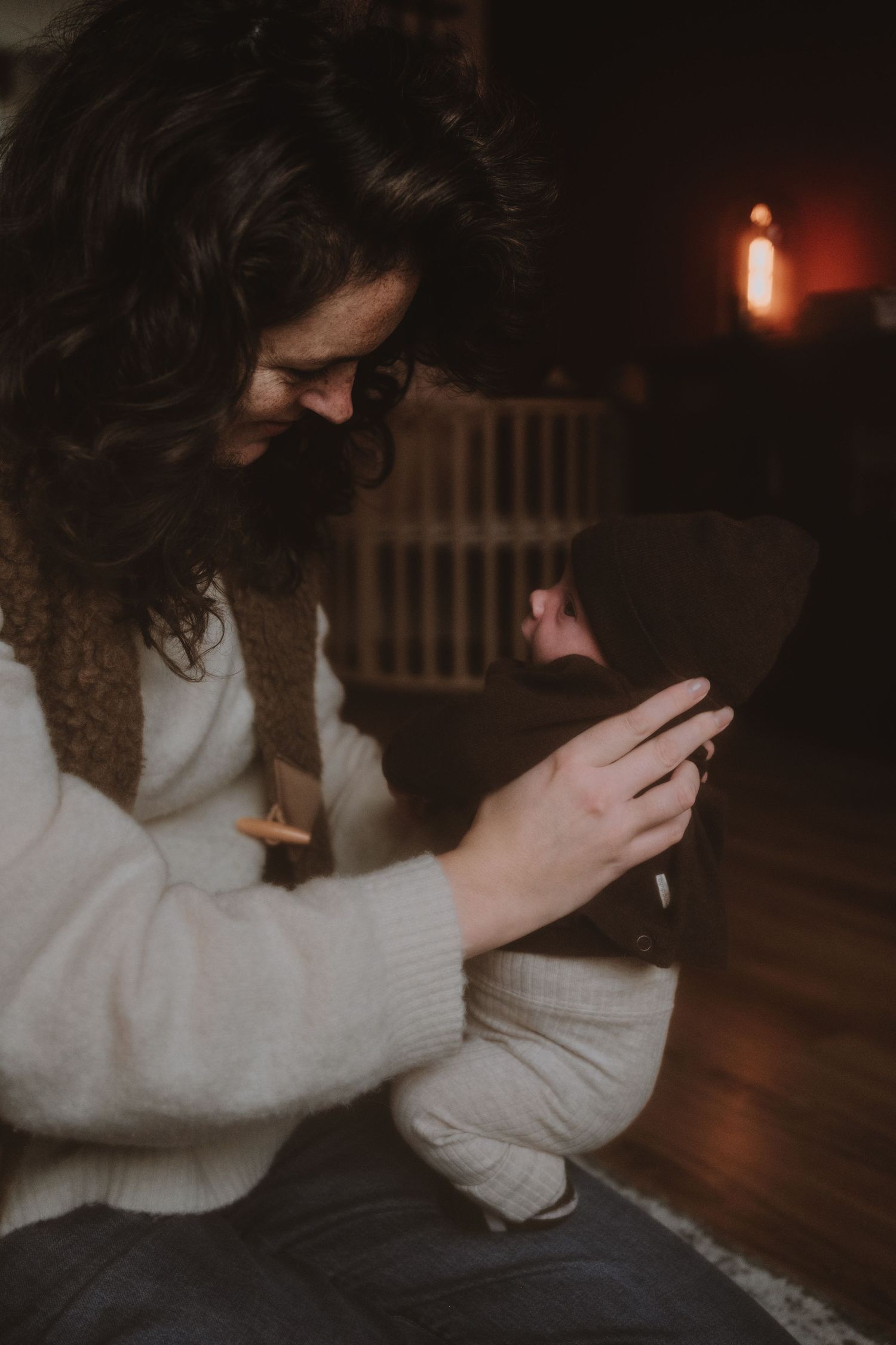 Mother in white sweater lovingly holds and examines her newborn baby in warm indoor setting.