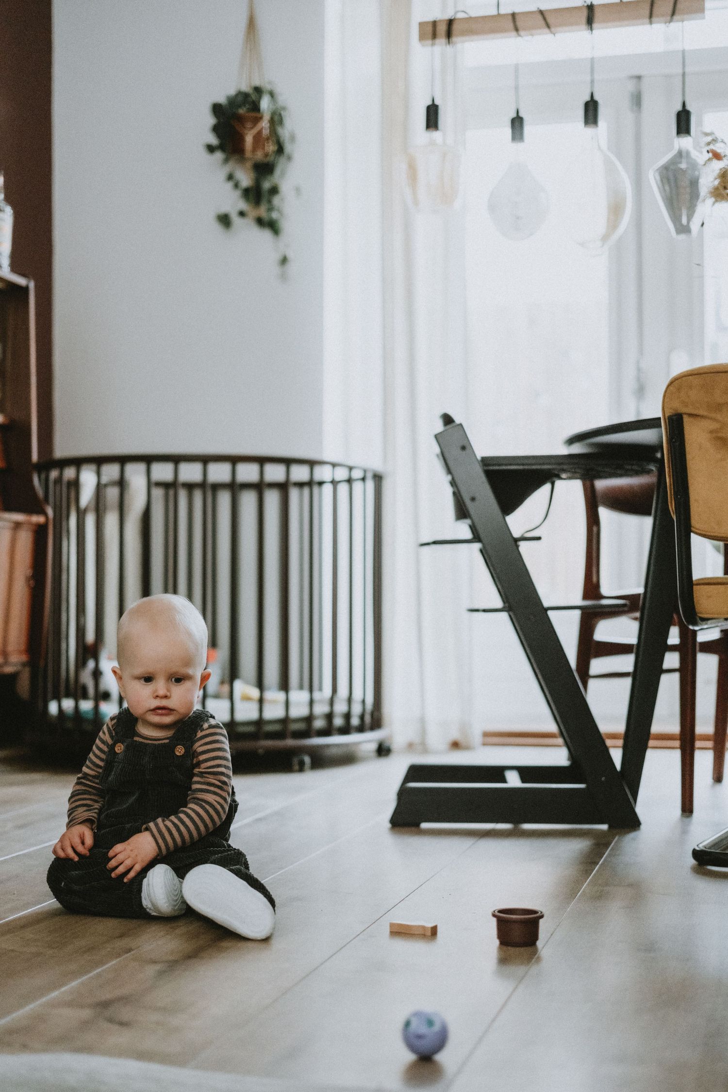 Cute baby in striped outfit sitting alone on wooden floor surrounded by colorful toys.