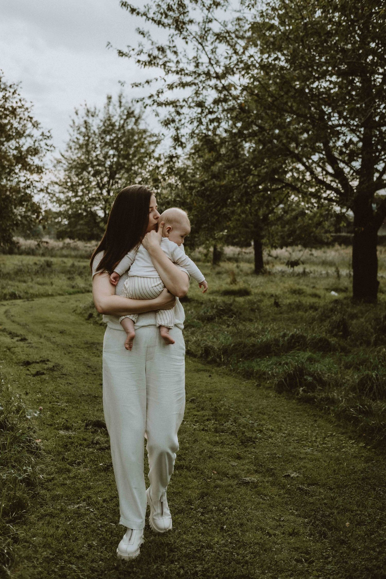 Young mother in white outfit kissing baby while standing in lush green orchard.