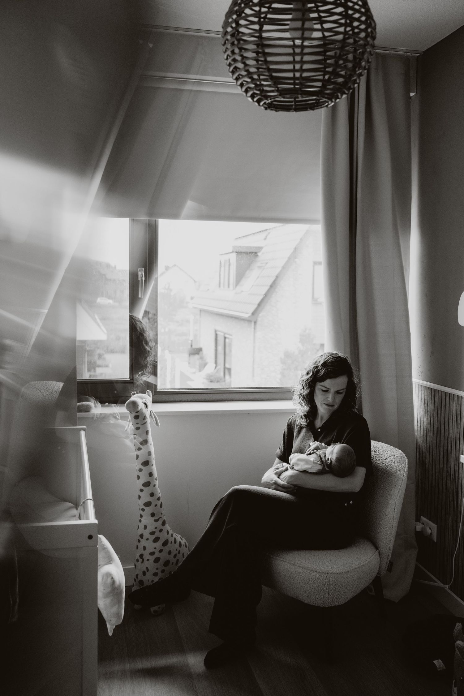 Black and white photo of mother breastfeeding newborn in nursery chair by window.