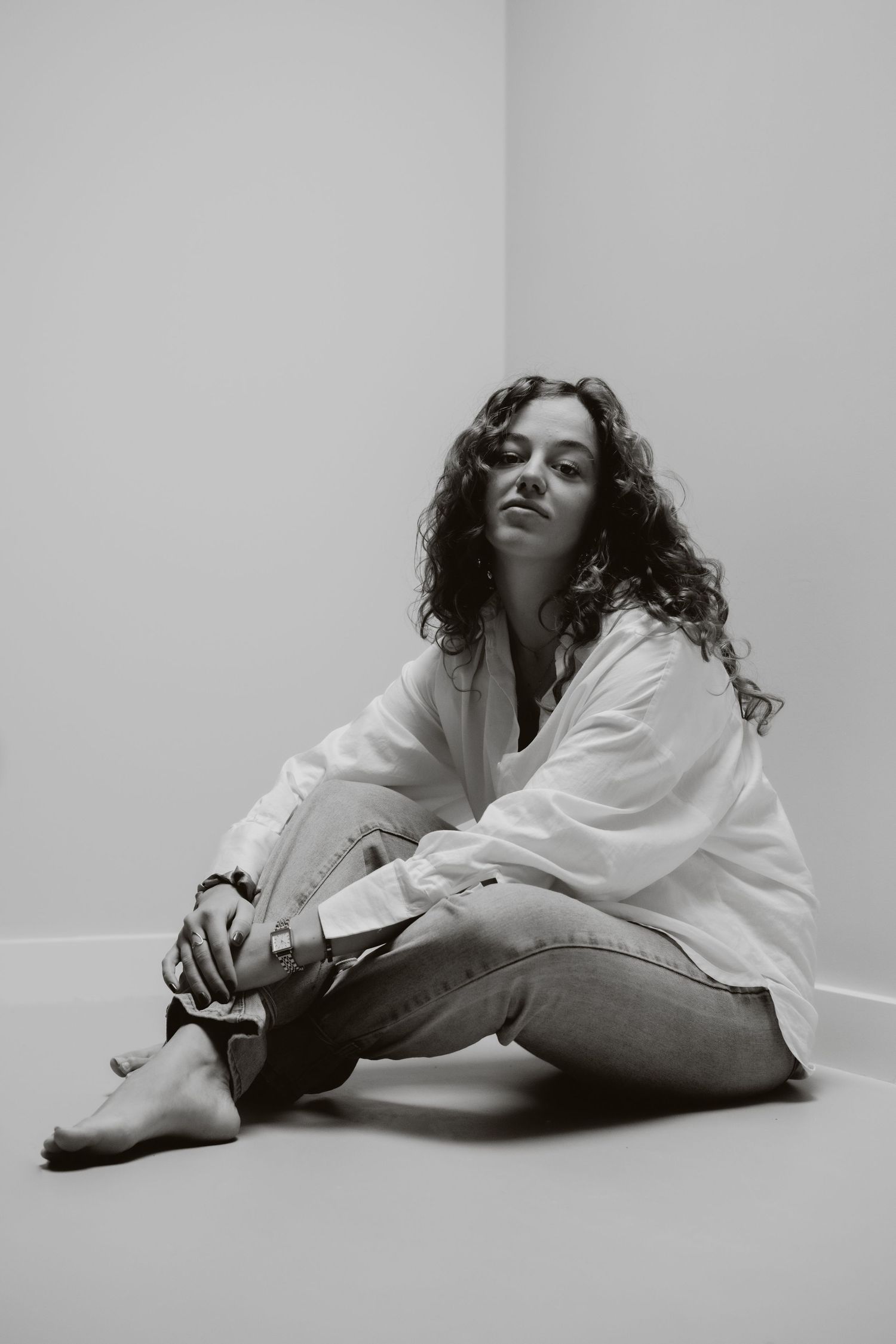 Black and white studio photo of a barefoot woman with curly hair sitting in a white shirt and jeans.