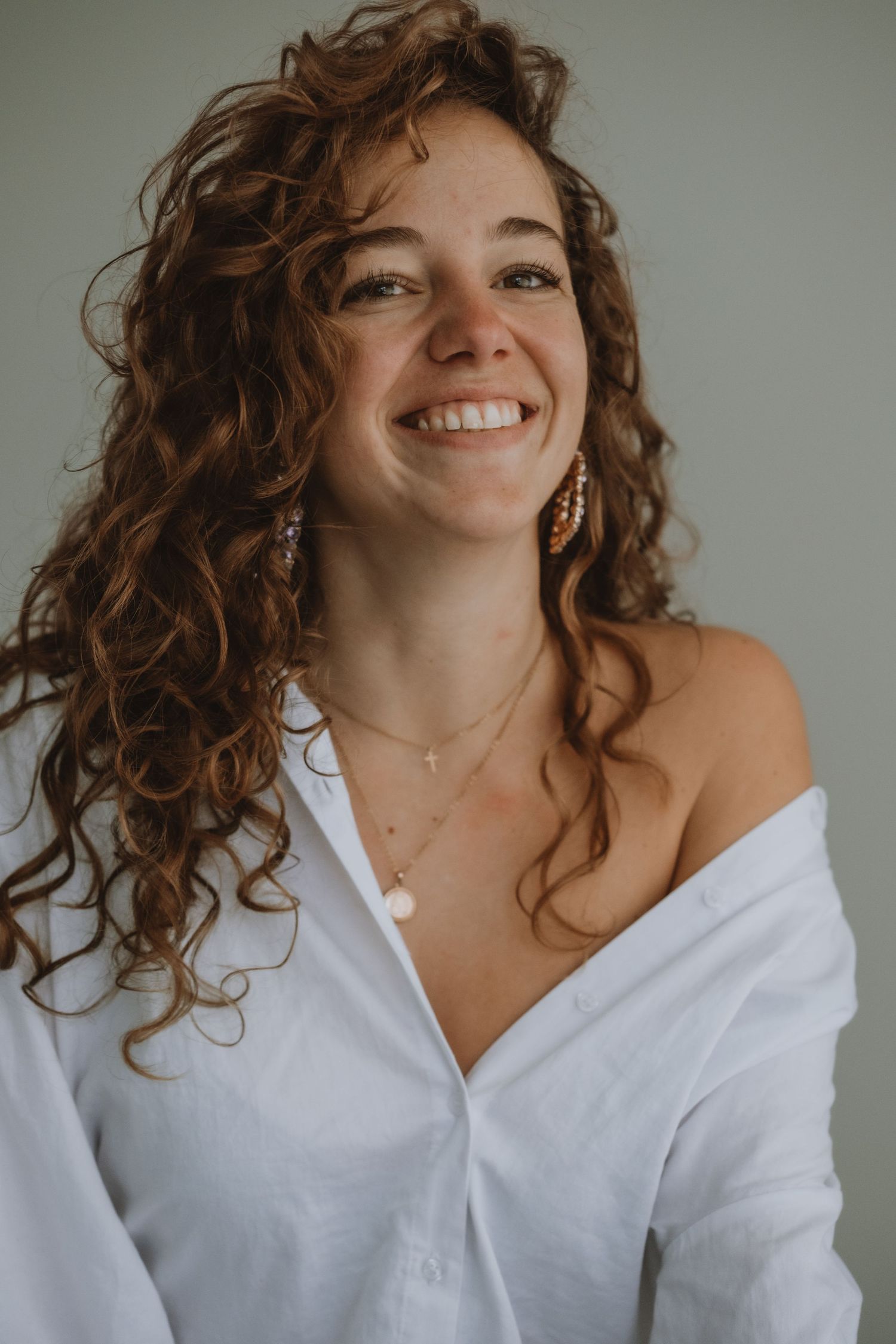 Cheerful woman with curly auburn hair in a white off-shoulder shirt wearing gold earrings and necklaces.