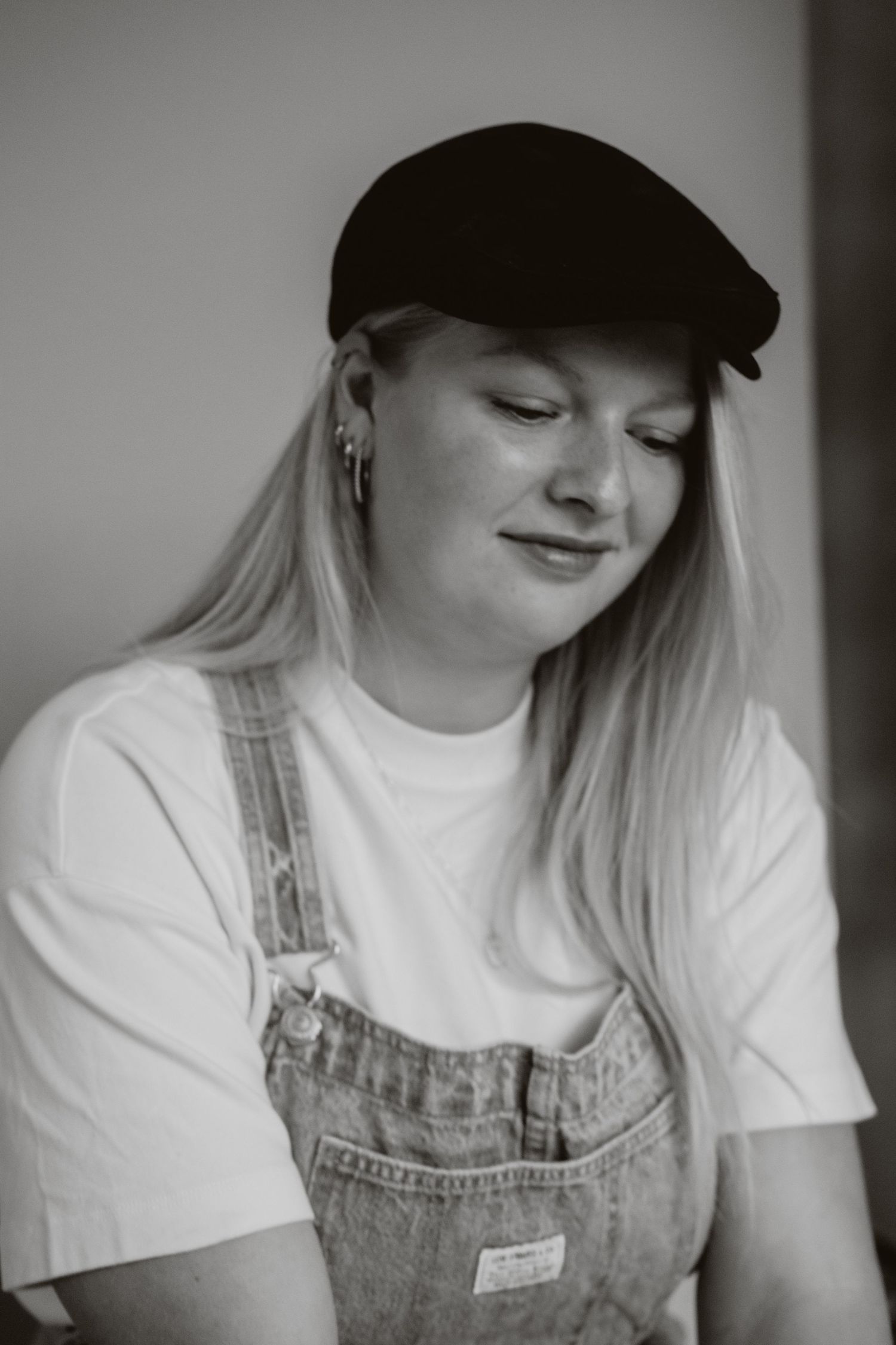 Black and white portrait of blonde woman in overalls and flat cap, smiling softly.