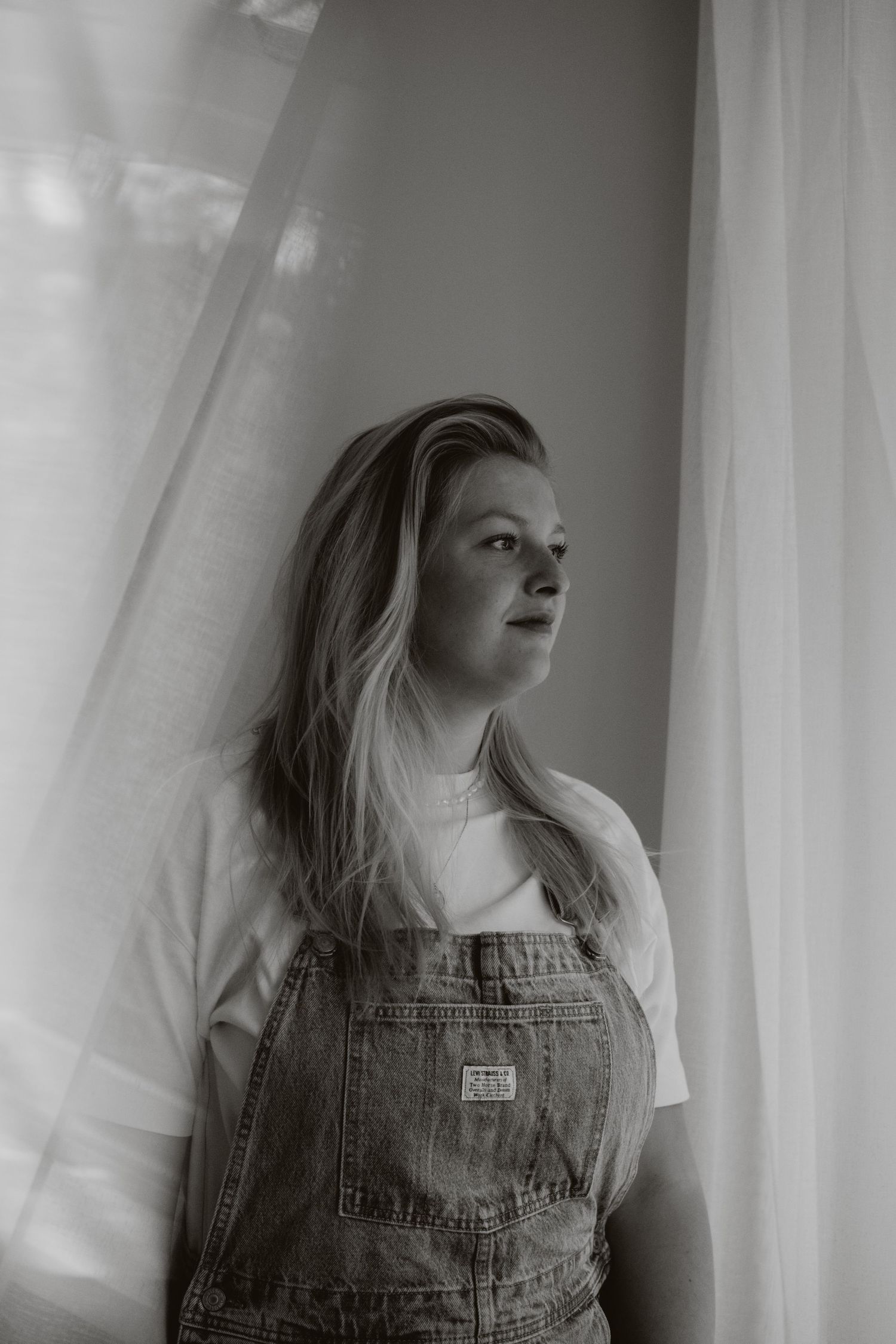 Black and white portrait of woman in overalls standing by sheer curtains.