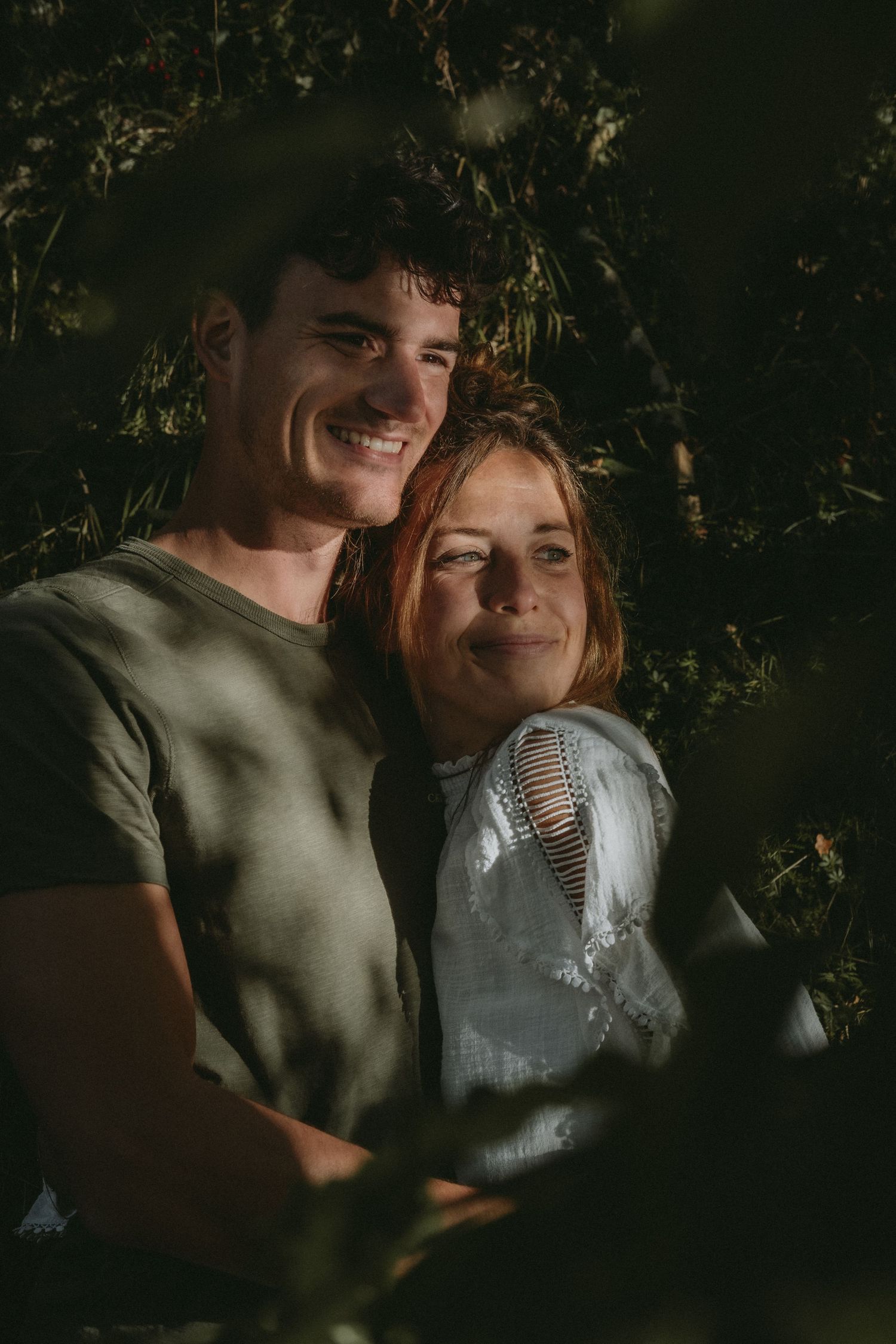 Smiling couple embracing outdoors surrounded by lush green foliage in dappled sunlight.