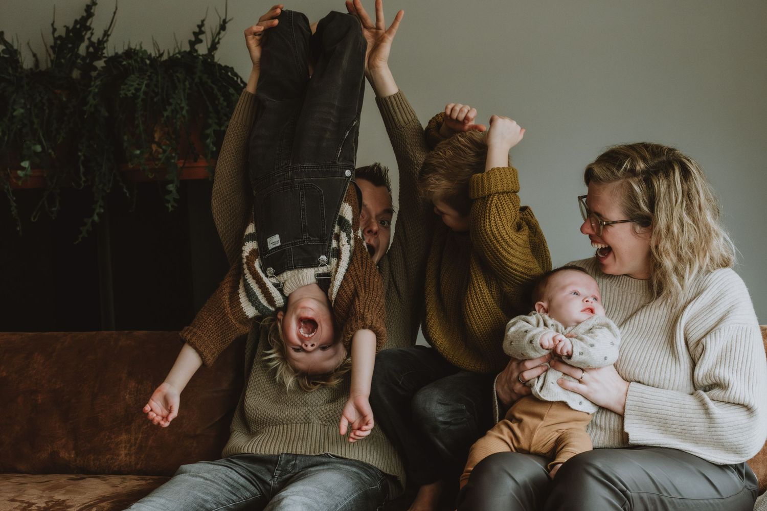 Lively family portrait with laughing parents, baby, and playful child hanging upside down on sofa.