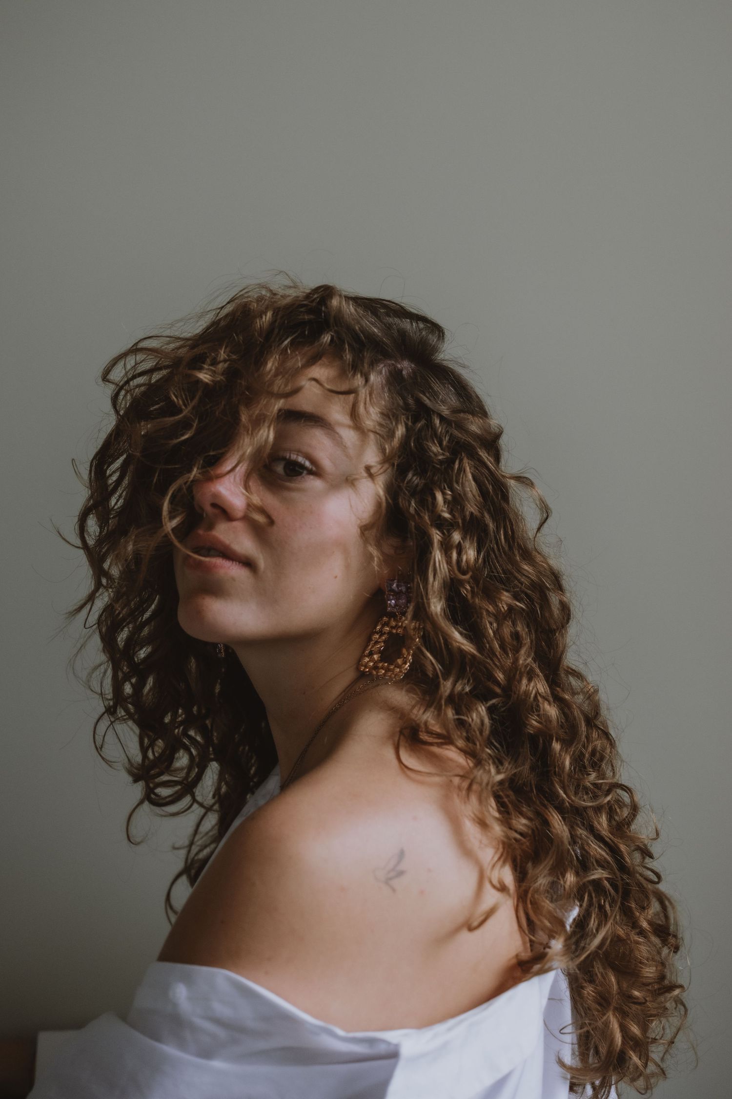 Woman with curly hair and large earrings wearing a white off-shoulder top with a small collarbone tattoo.