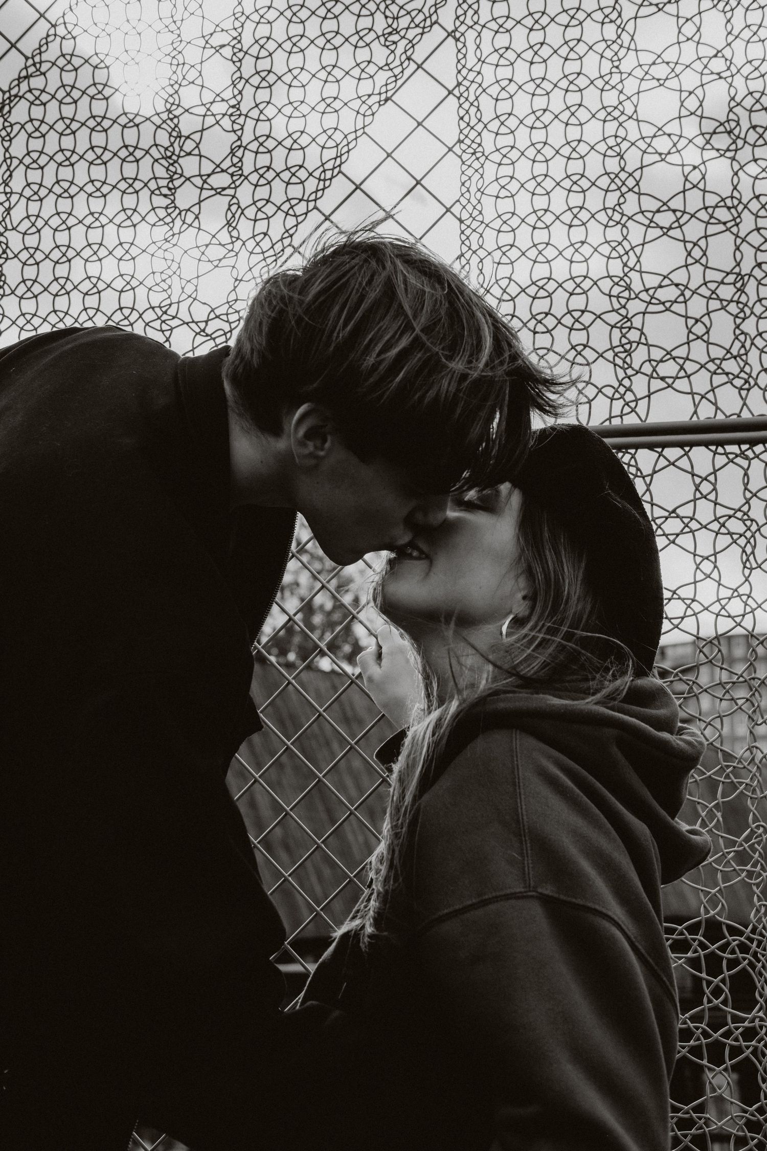 Black and white close-up of a young couple sharing a kiss against a chain-link fence outdoors.