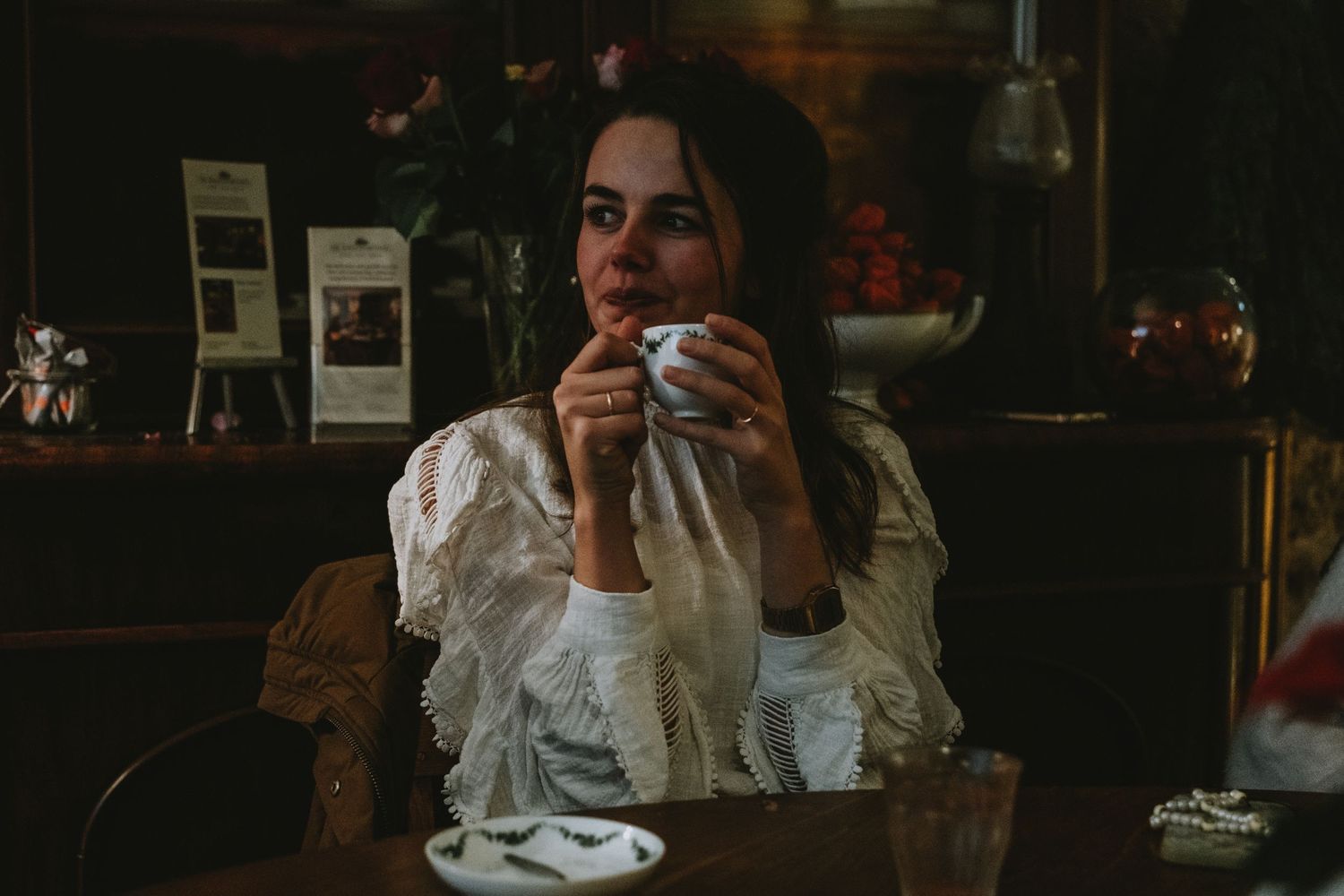 Young woman in vintage lace dress sipping from a cup in a dimly lit cozy cafe setting.