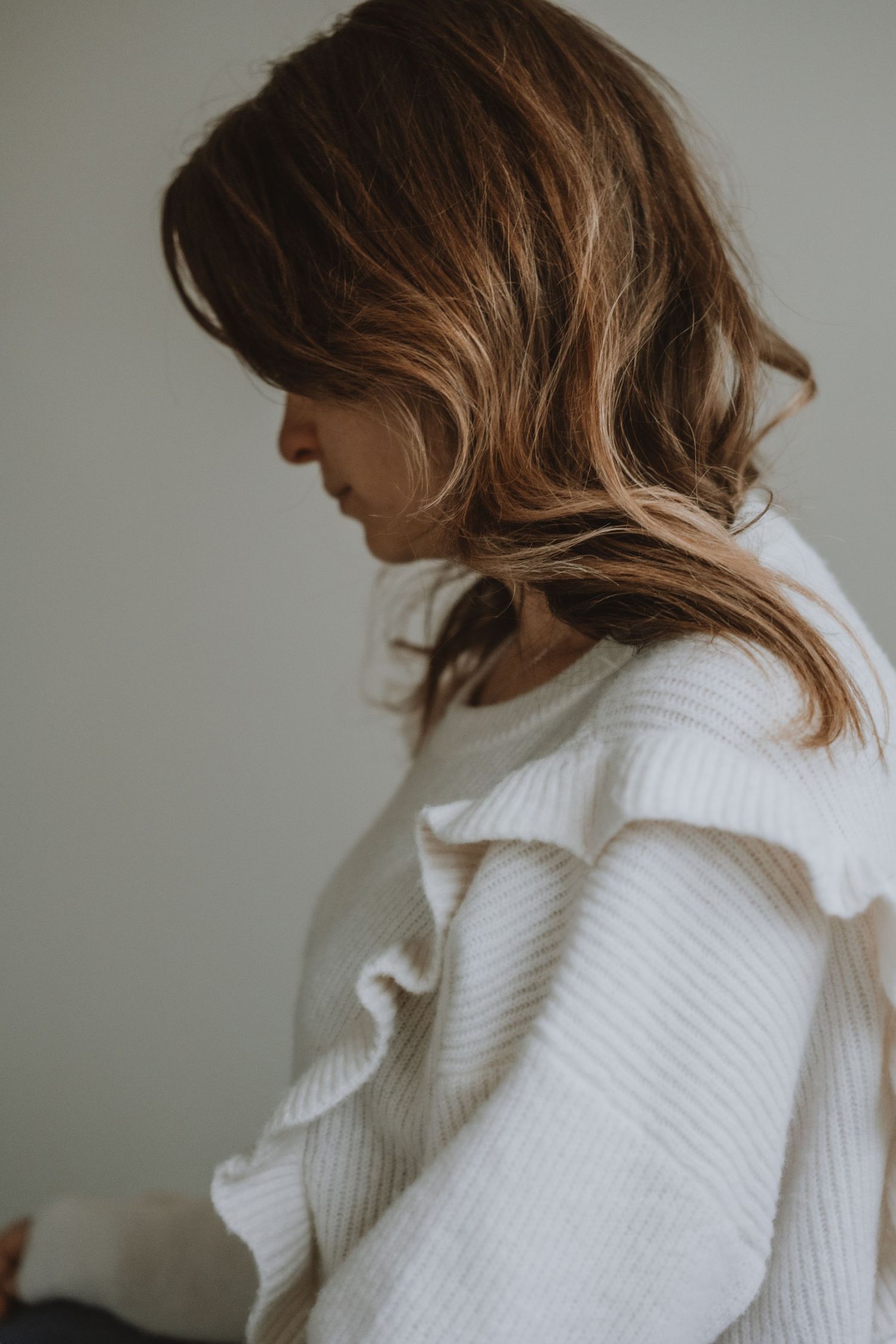 Side profile of a woman with brown wavy hair wearing a white ruffled sweater.