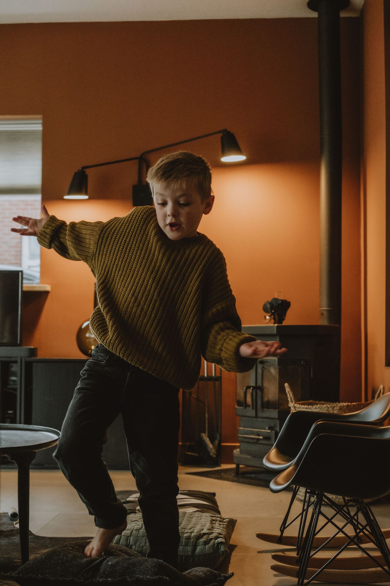 Young boy in olive sweater playfully dancing in a warm orange living room.