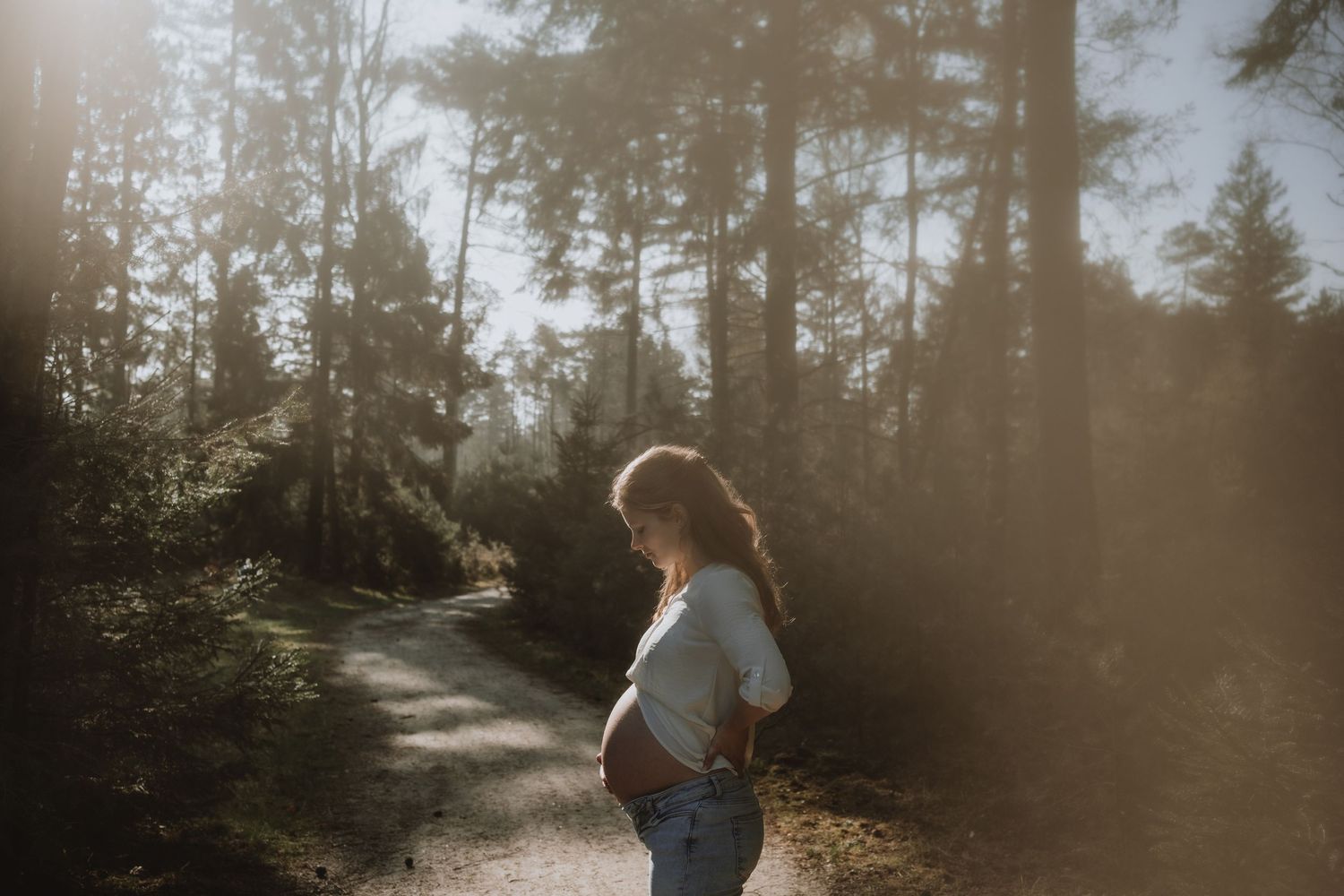 Pregnant woman standing alone on sunlit forest path, cradling baby bump, soft light filtering through trees.