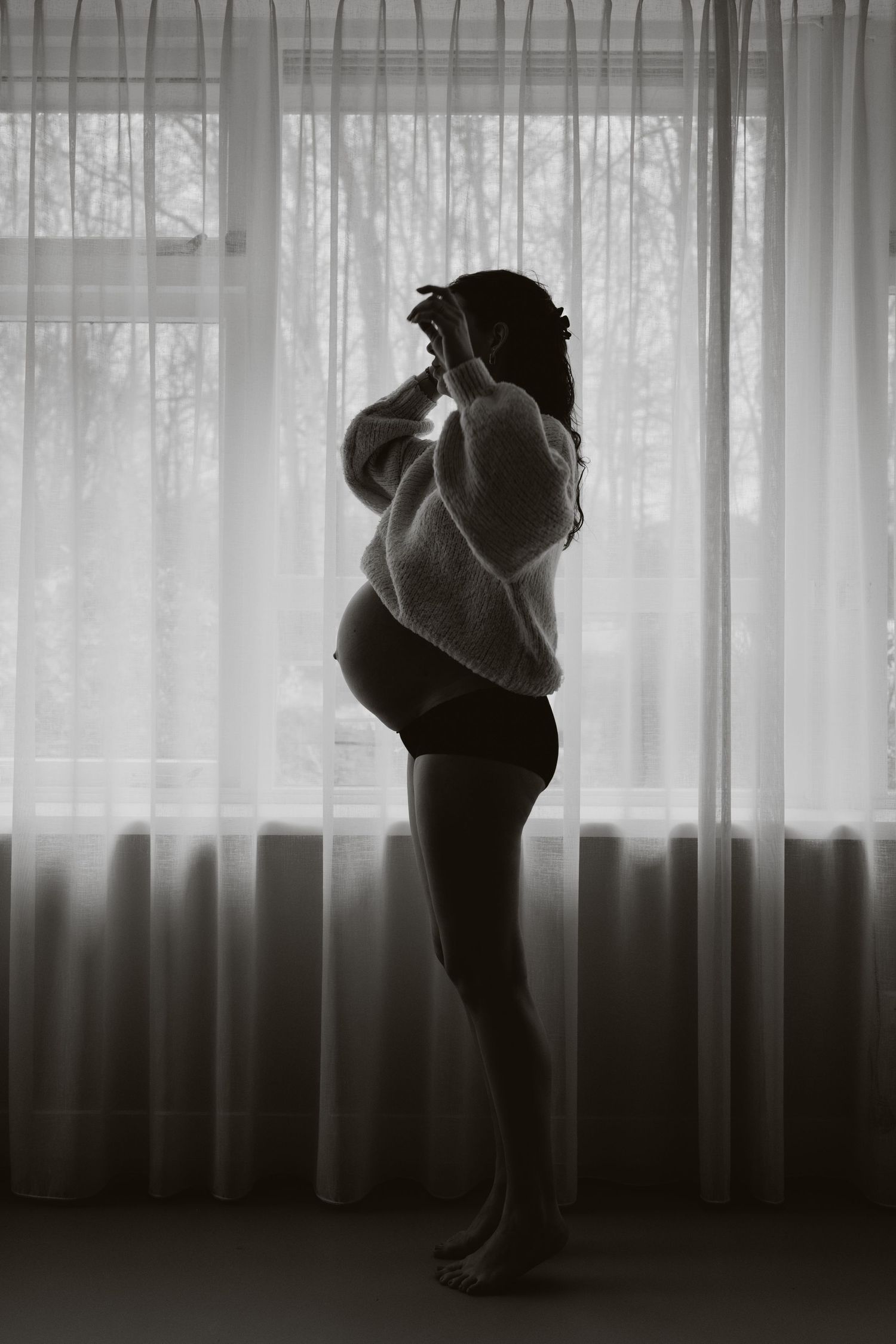 Silhouette of pregnant woman in fuzzy sweater standing before sheer curtained window.