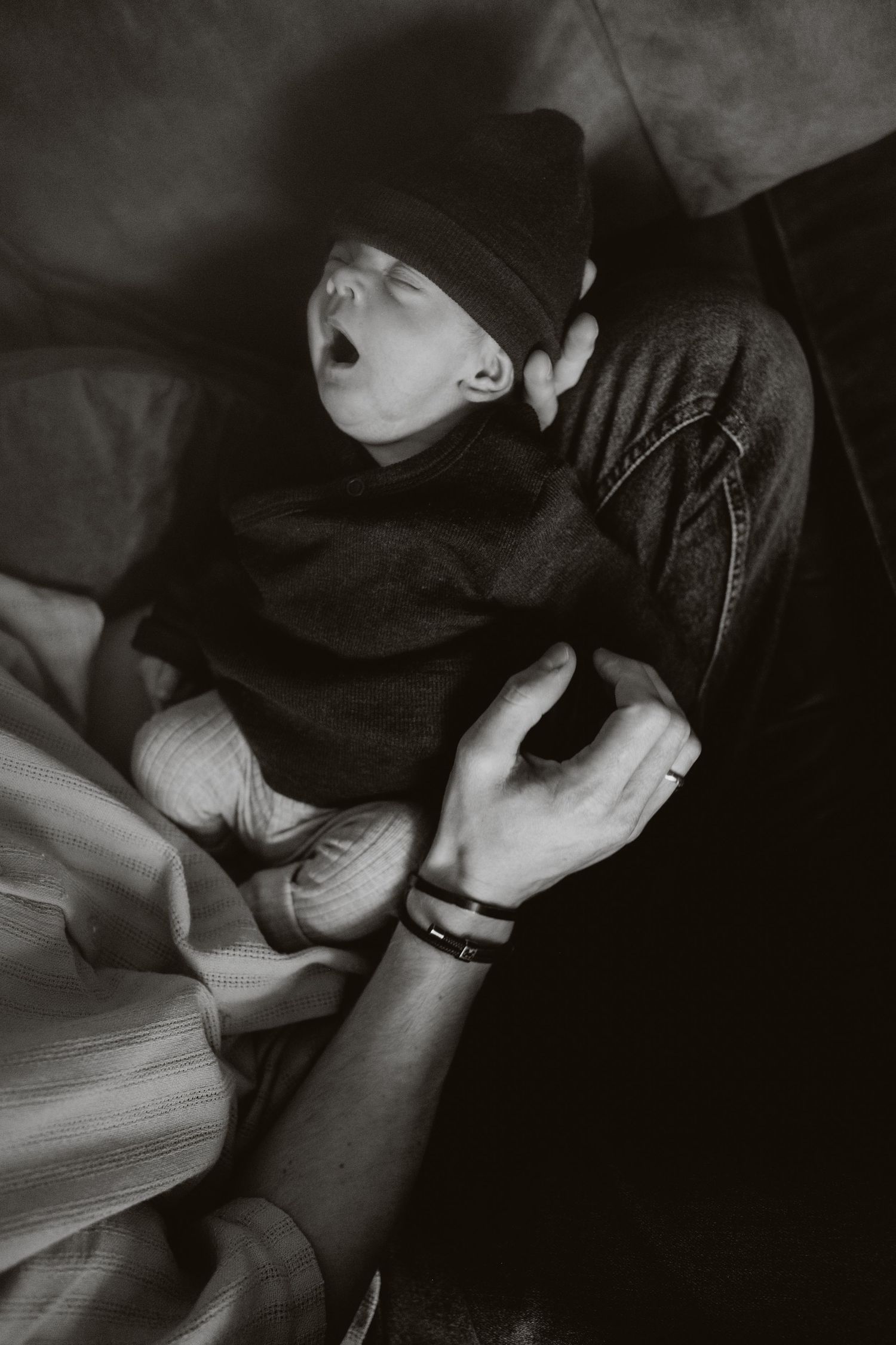 Black and white photo of yawning newborn baby in knit hat resting on parent's lap.