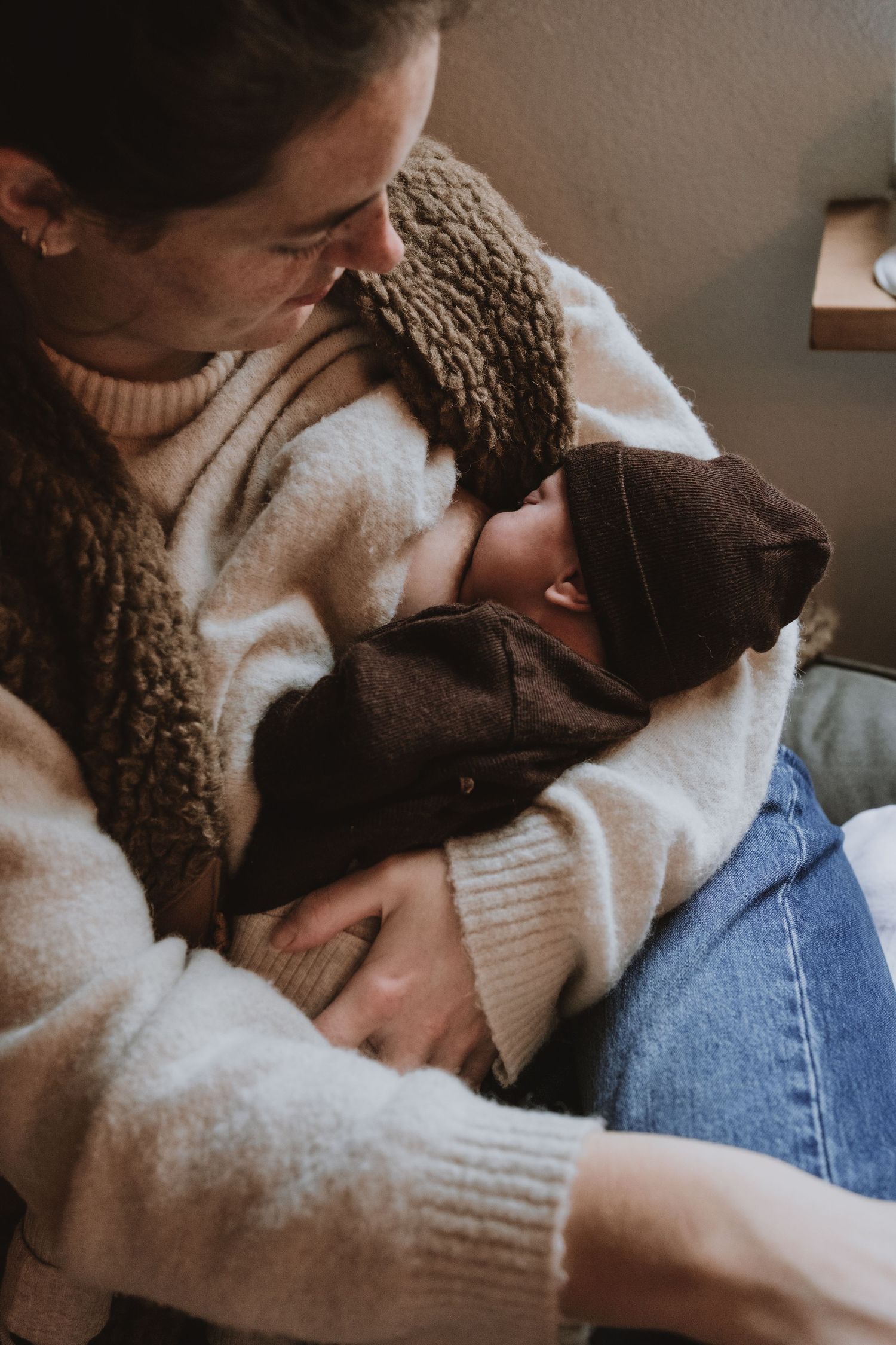 Mother in cozy white sweater smiles lovingly while holding sleeping newborn in dark knit hat.