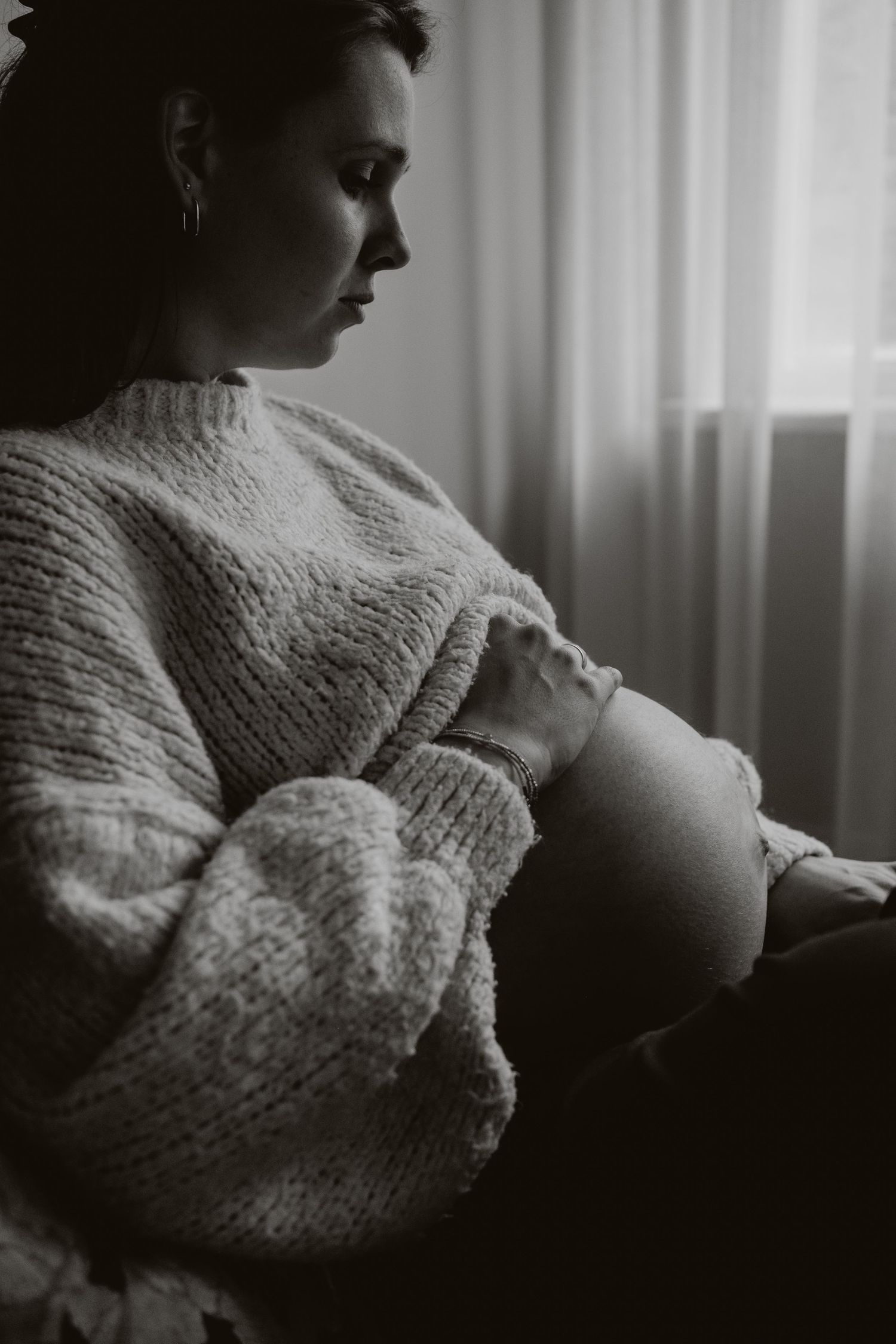 Black and white photo of pregnant woman in knit sweater cradling baby bump while sitting by window.