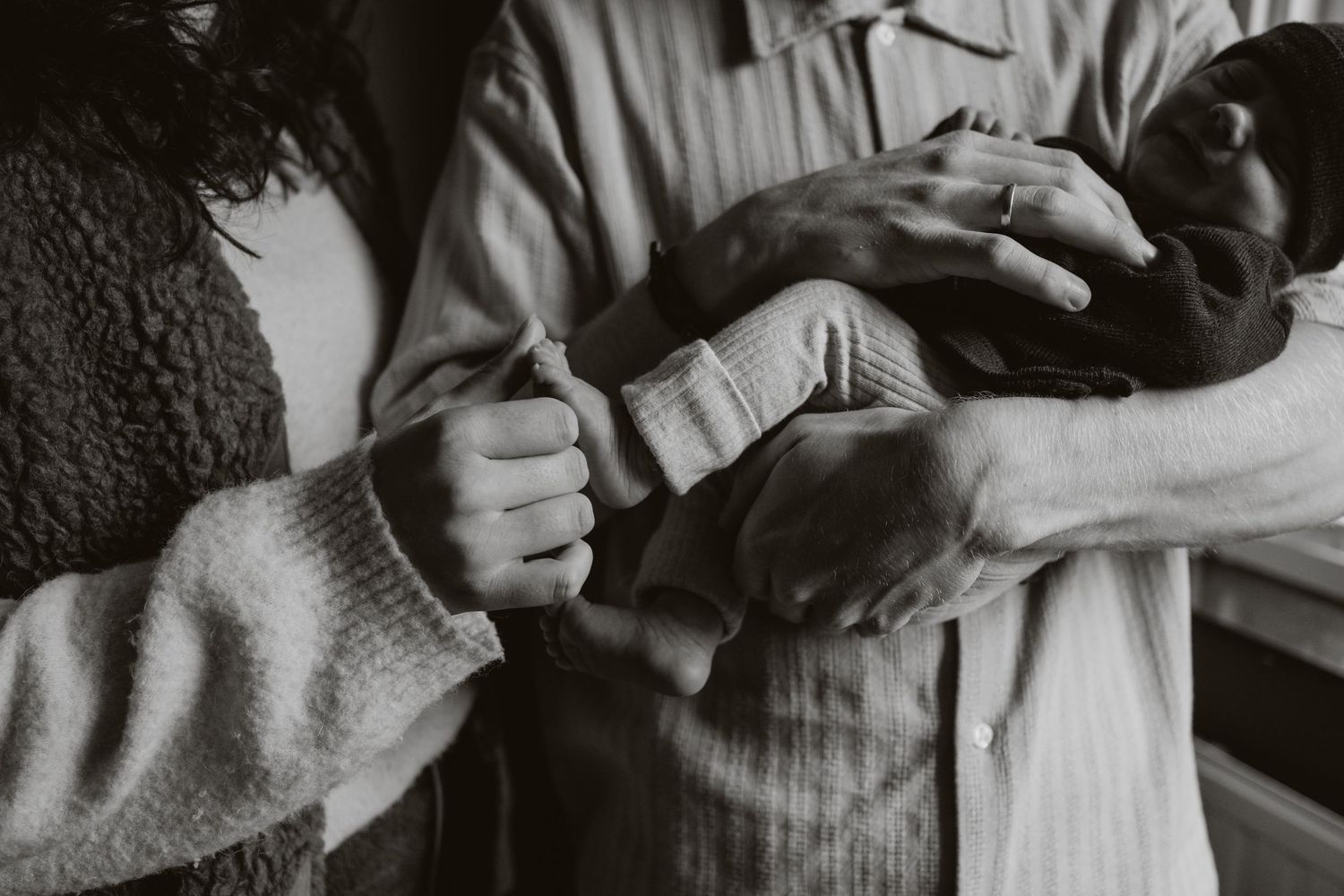 Black and white close-up of new parents hands gently holding their swaddled newborn baby.
