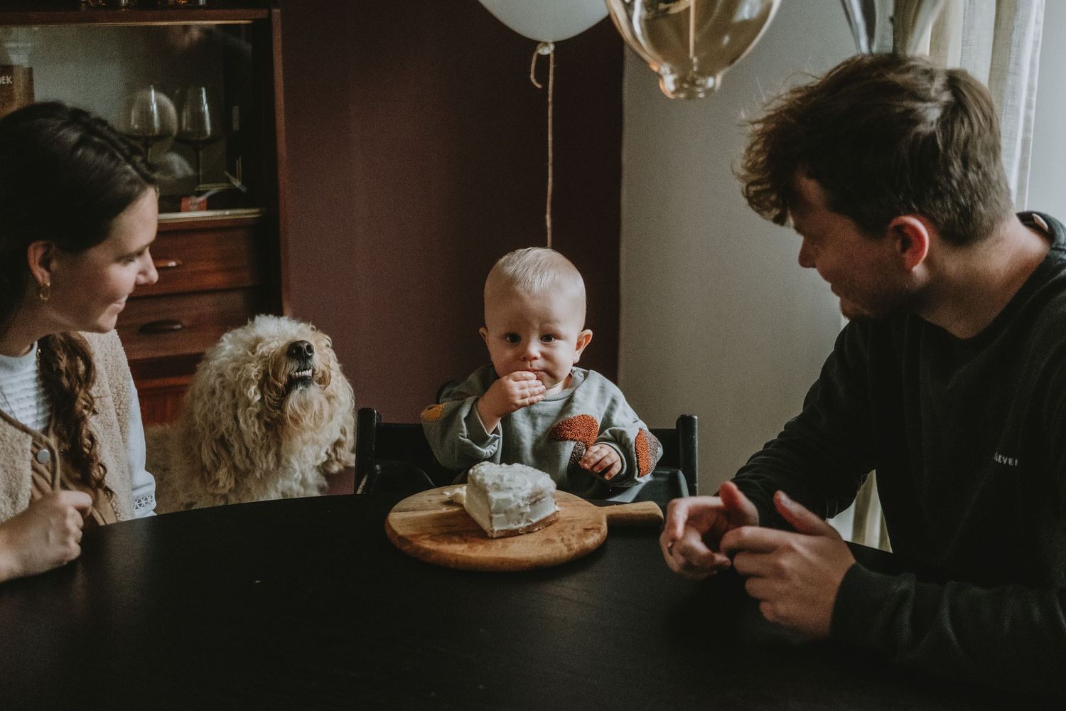 Family celebrating baby first birthday with cake at dining table with fluffy dog nearby.