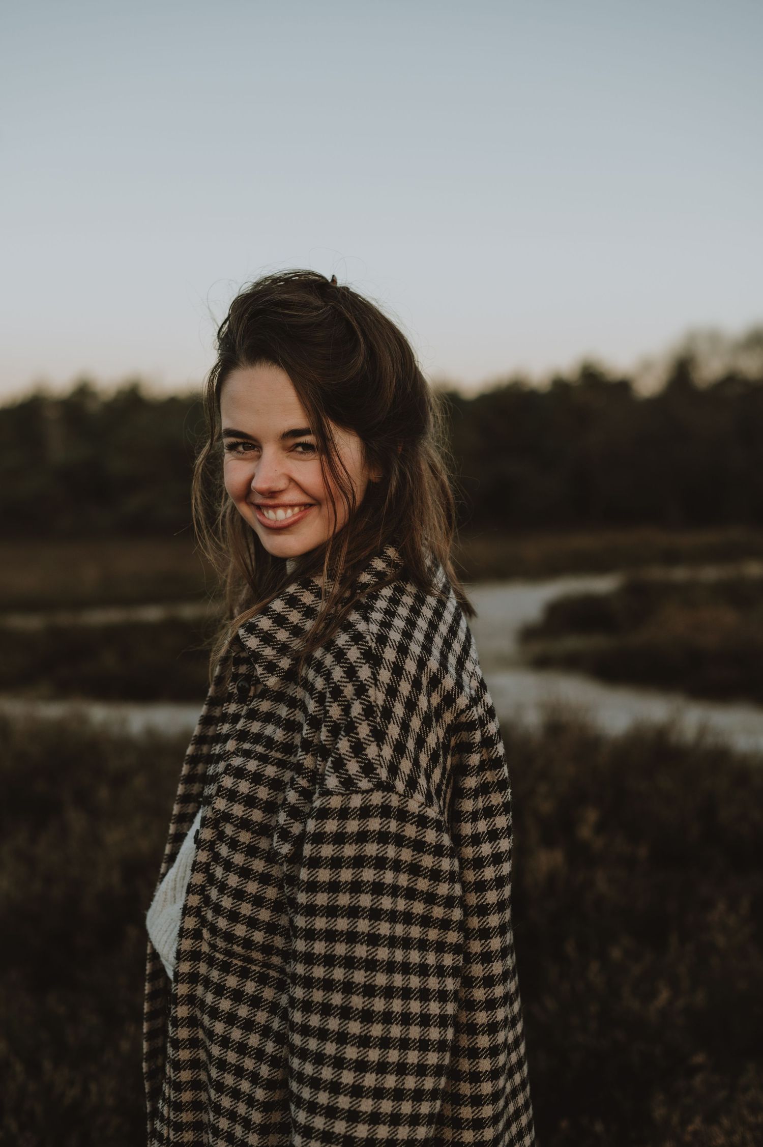 Young woman smiling over shoulder in plaid coat with autumn landscape background.