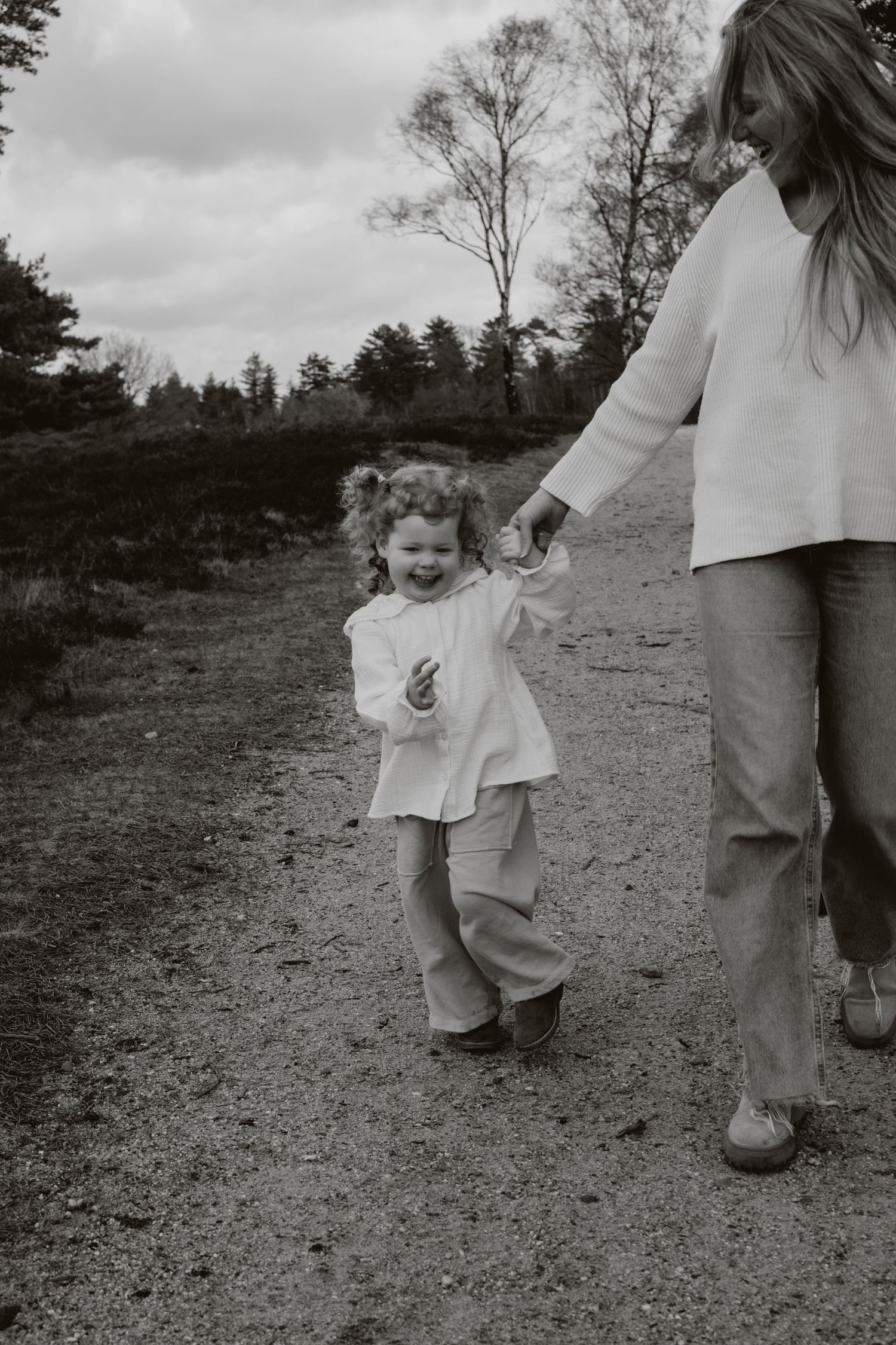 A happy curly-haired toddler walks hand-in-hand with her mother on a dirt path.