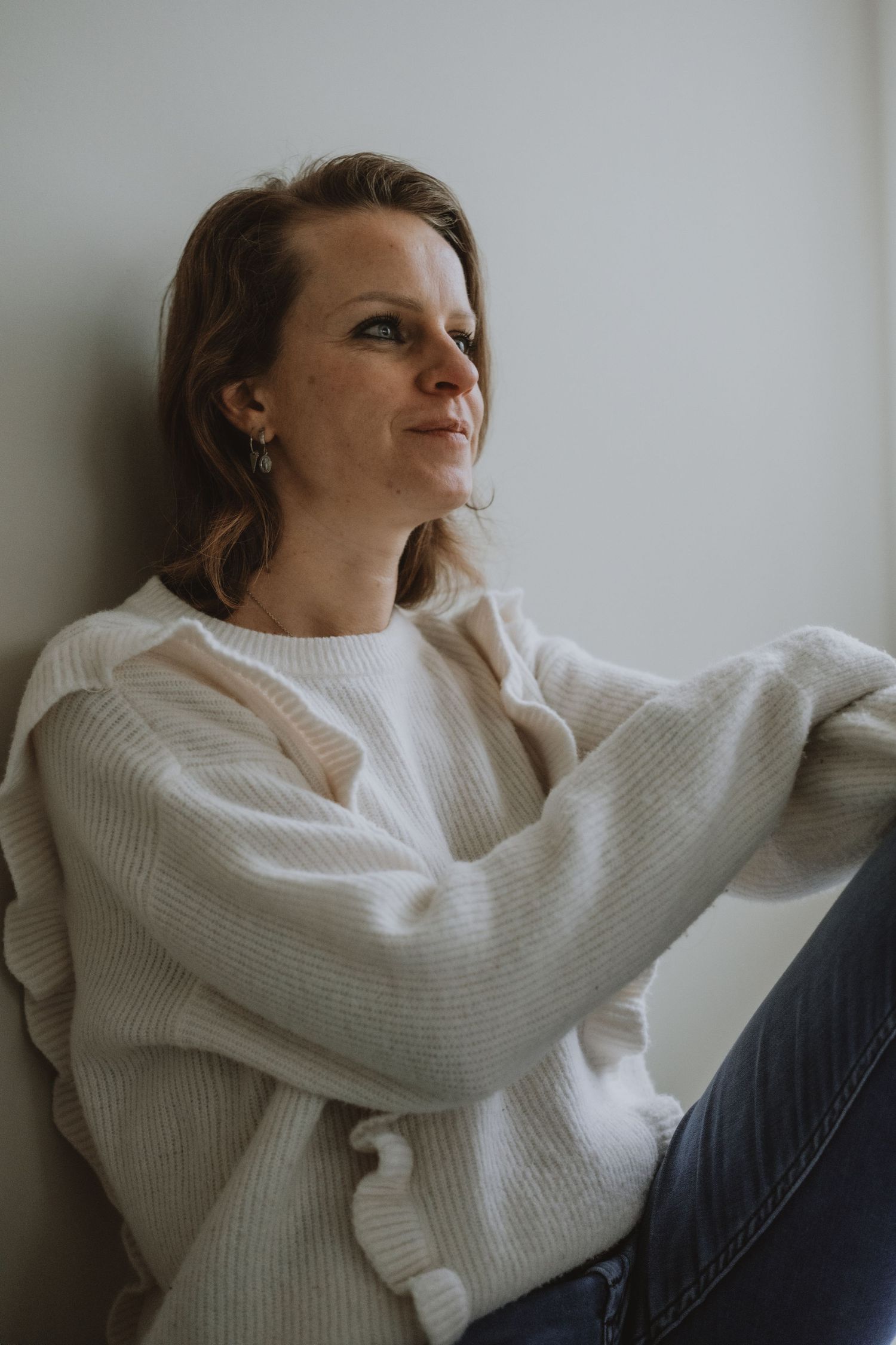 Woman in cream knit sweater and jeans sitting against a white wall smiling softly.