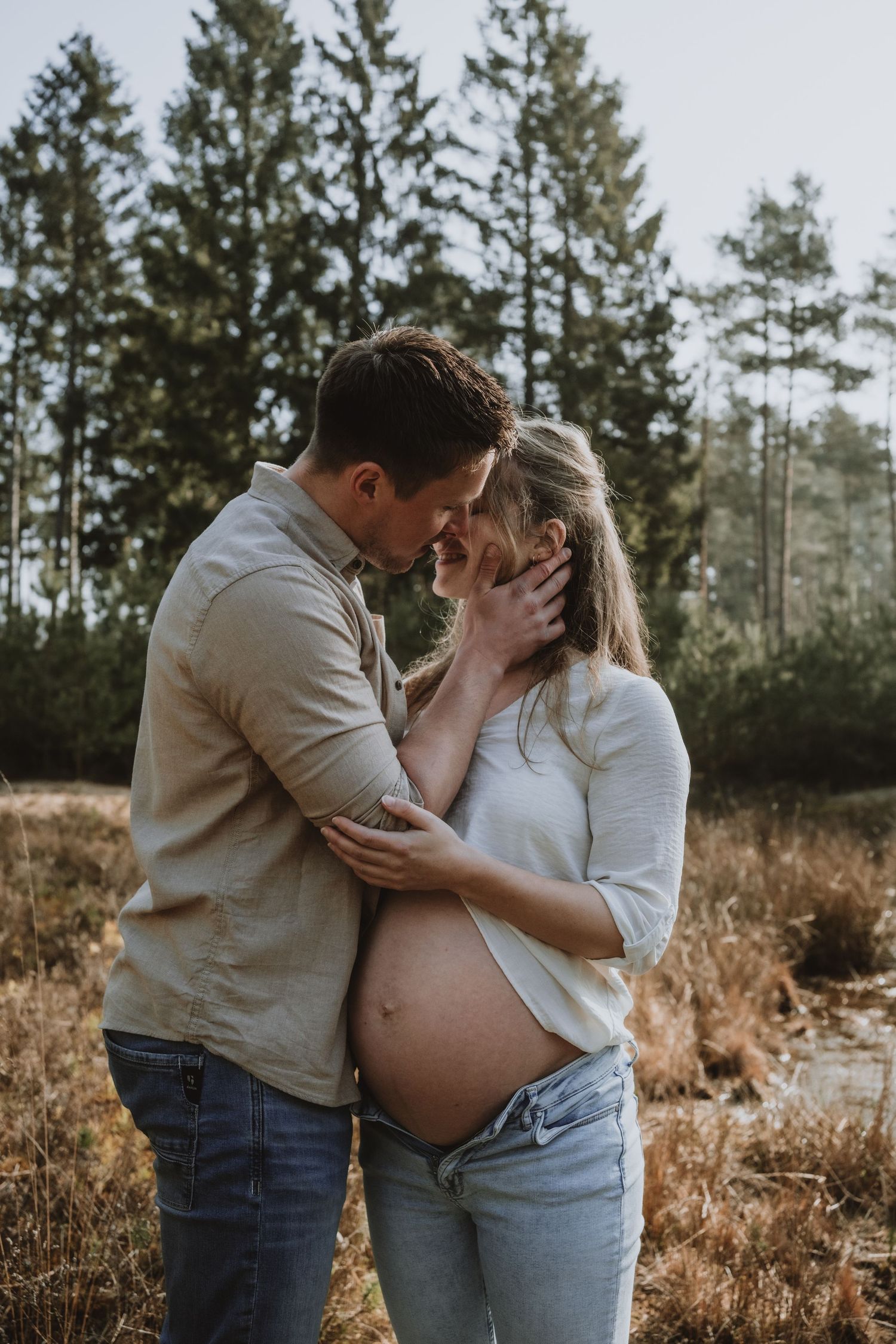 Couple sharing intimate moment in sunny field, pregnant woman's bare belly visible, pine forest background.