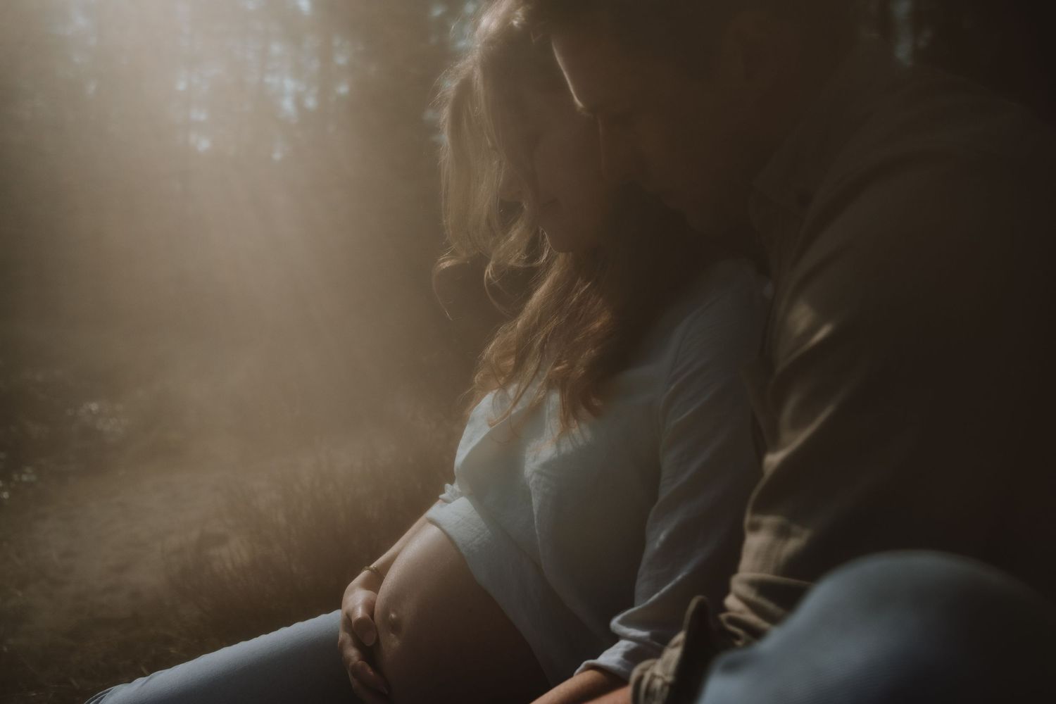 Pregnant woman sitting in soft sunlight in forest, cradling belly, warm golden rays filtering through trees.