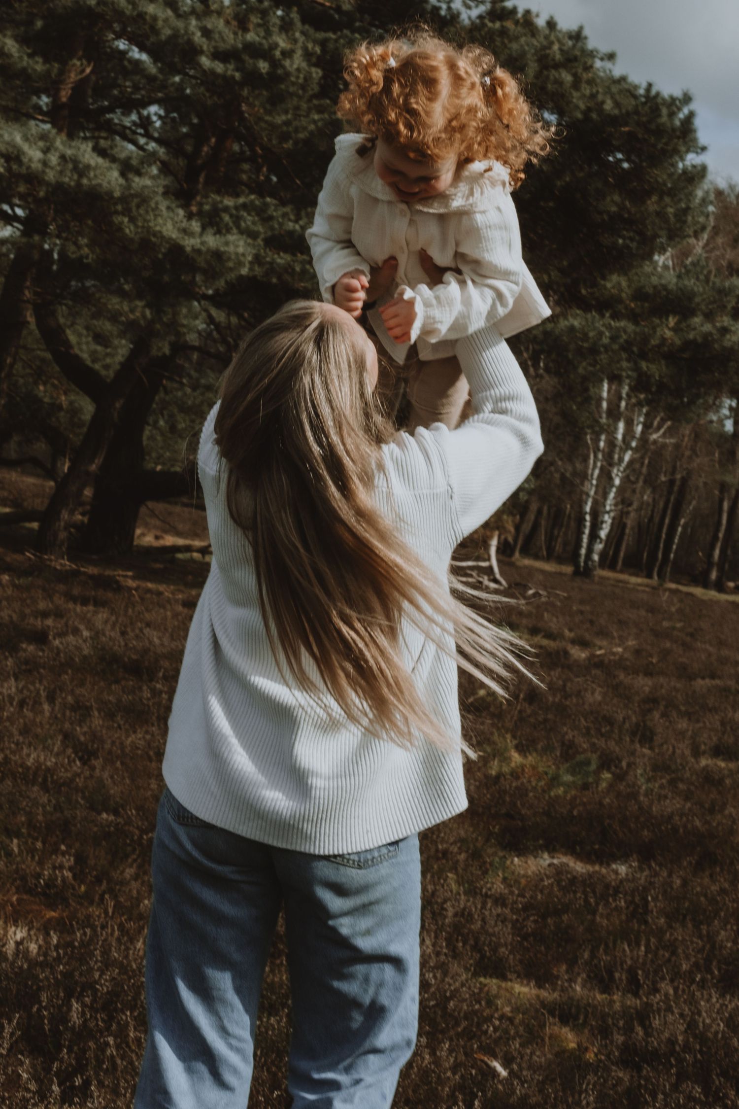 A mother lifts her curly-haired toddler joyfully in an autumn forest setting.