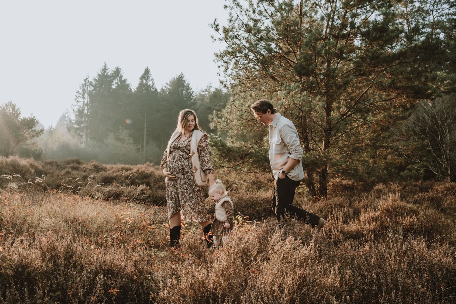 A pregnant couple walks with their toddler through a sunlit heathland field among trees.