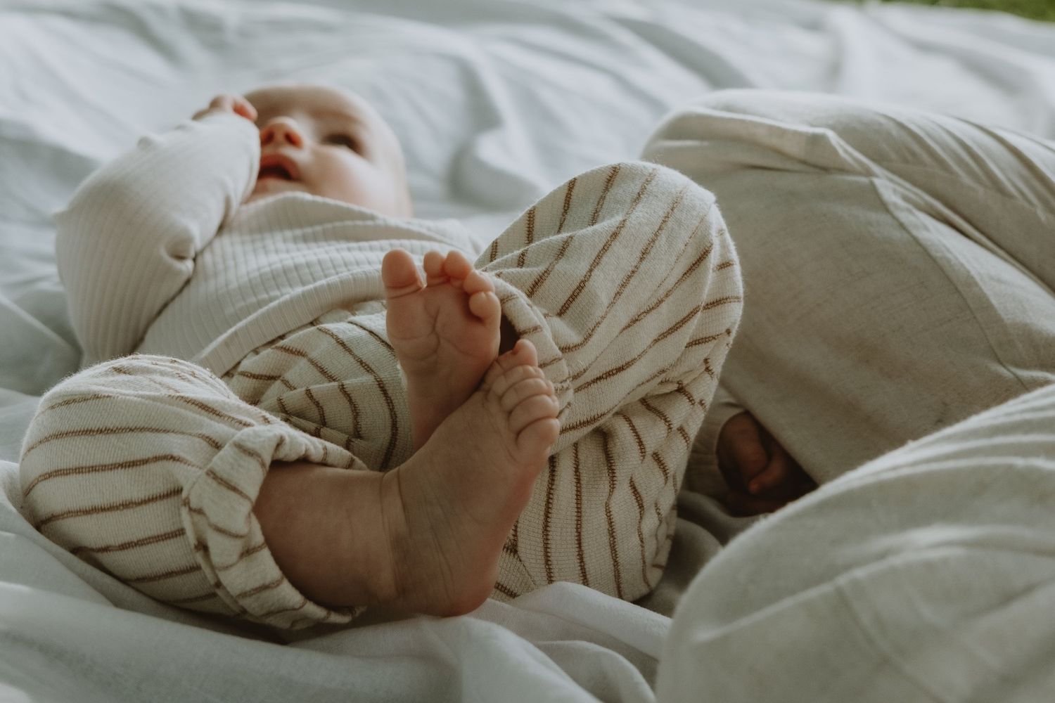 Peaceful baby lying on white bedding wearing striped outfit with tiny bare feet visible.