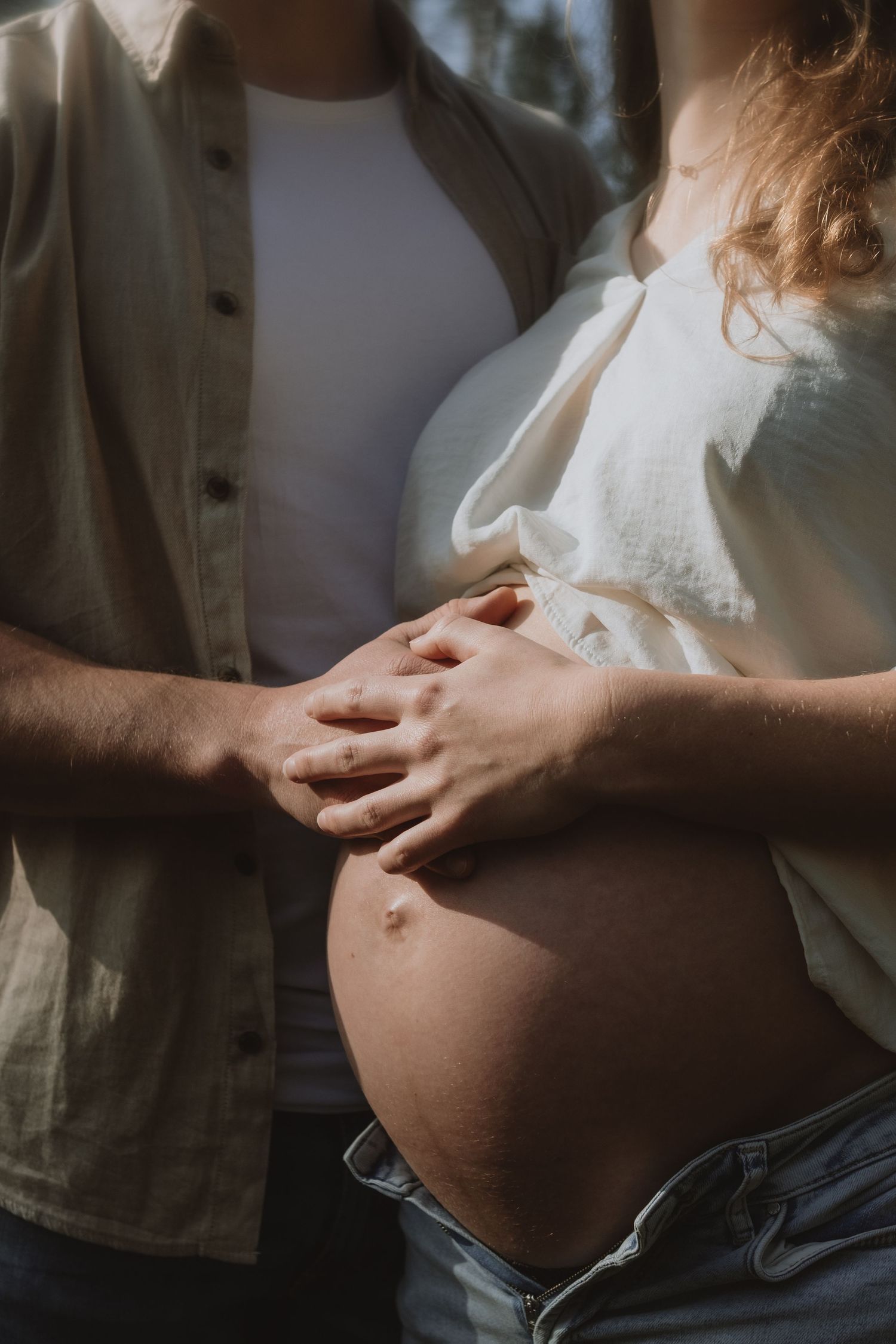 Couple tenderly holding pregnant belly together outdoors in warm natural sunlight.