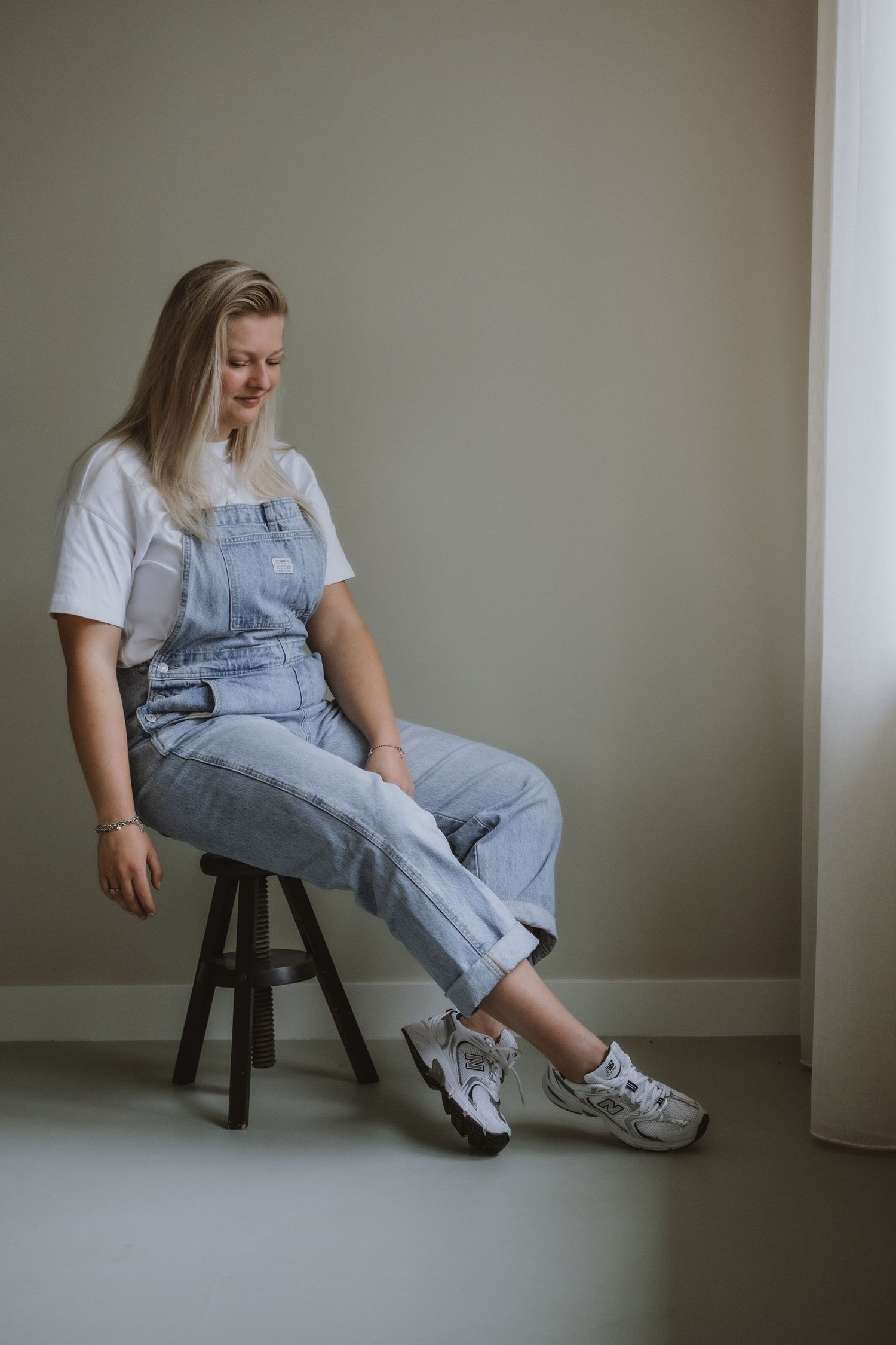 Young woman in denim overalls and white tee sits on wooden stool against neutral wall.